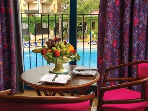 A dining table with a fruit centerpiece, a chair, and a view of a pool and buildings. - Hotel Caesar Palace (Giardini Naxos)