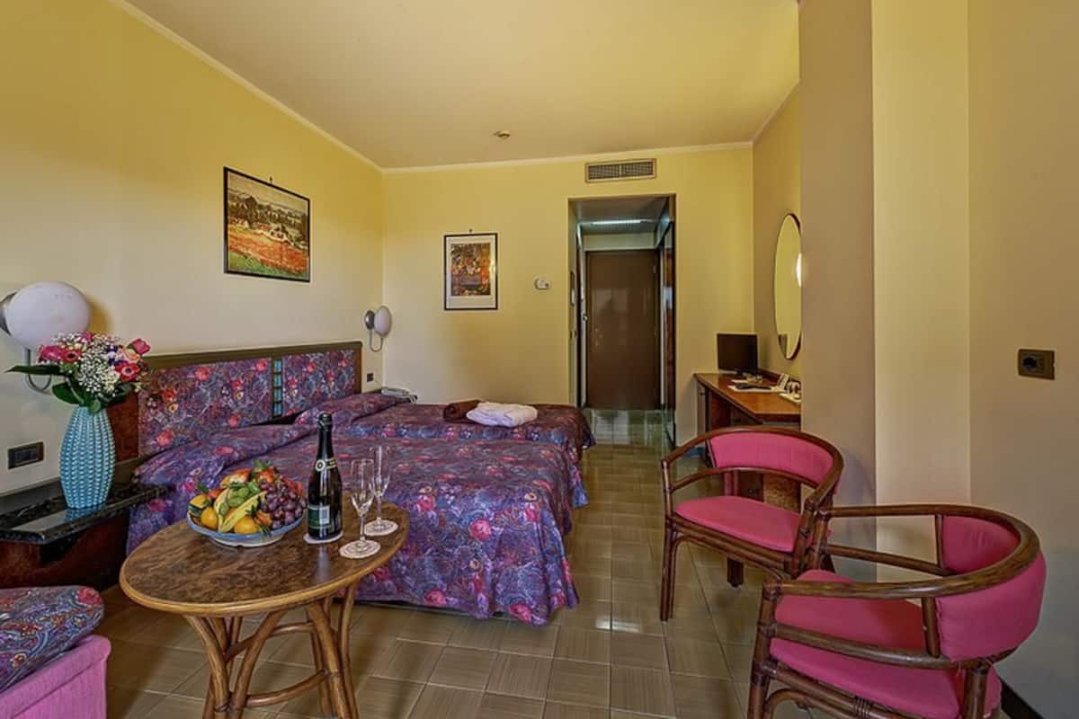 Une chambre d’hôtel comprenant un lit, un bureau, une chaise, une table avec un panier de fruits et des boissons, et un vase avec des fleurs.