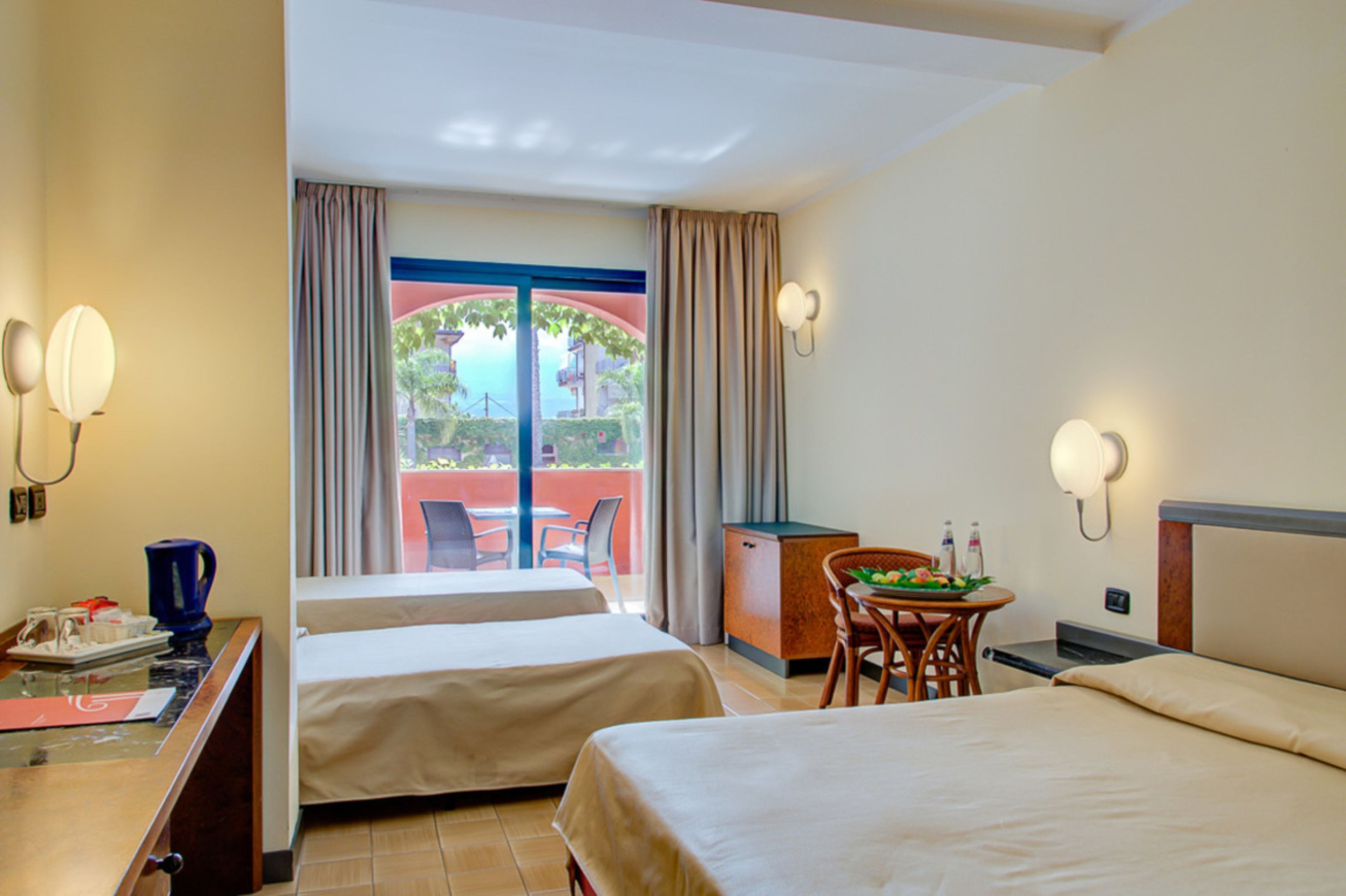 A hotel room with two beds, a desk with a chair, a small table with a plant, and a view of a red building and greenery outside.