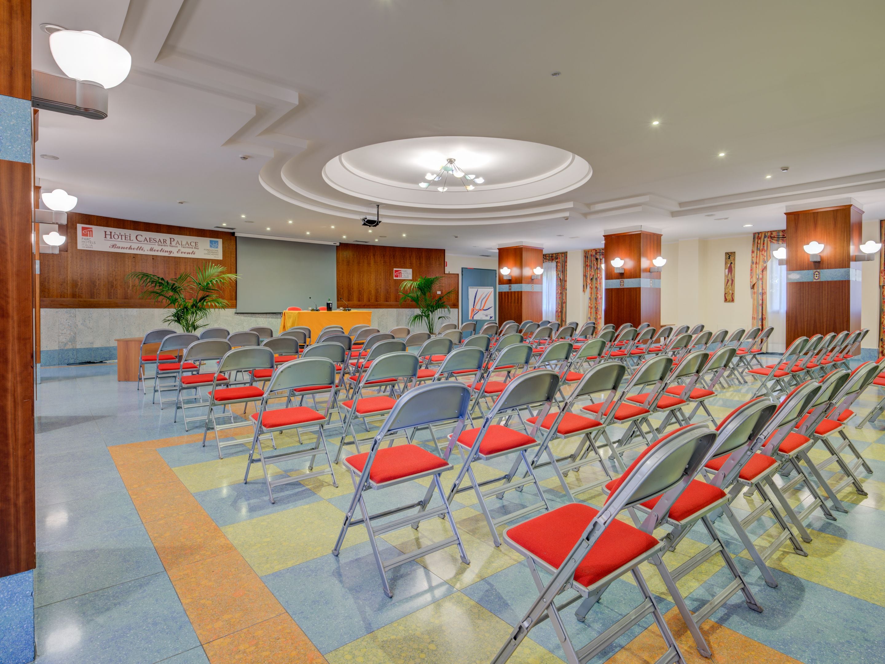 A hotel conference room with rows of foldable chairs, a podium, and a large screen.