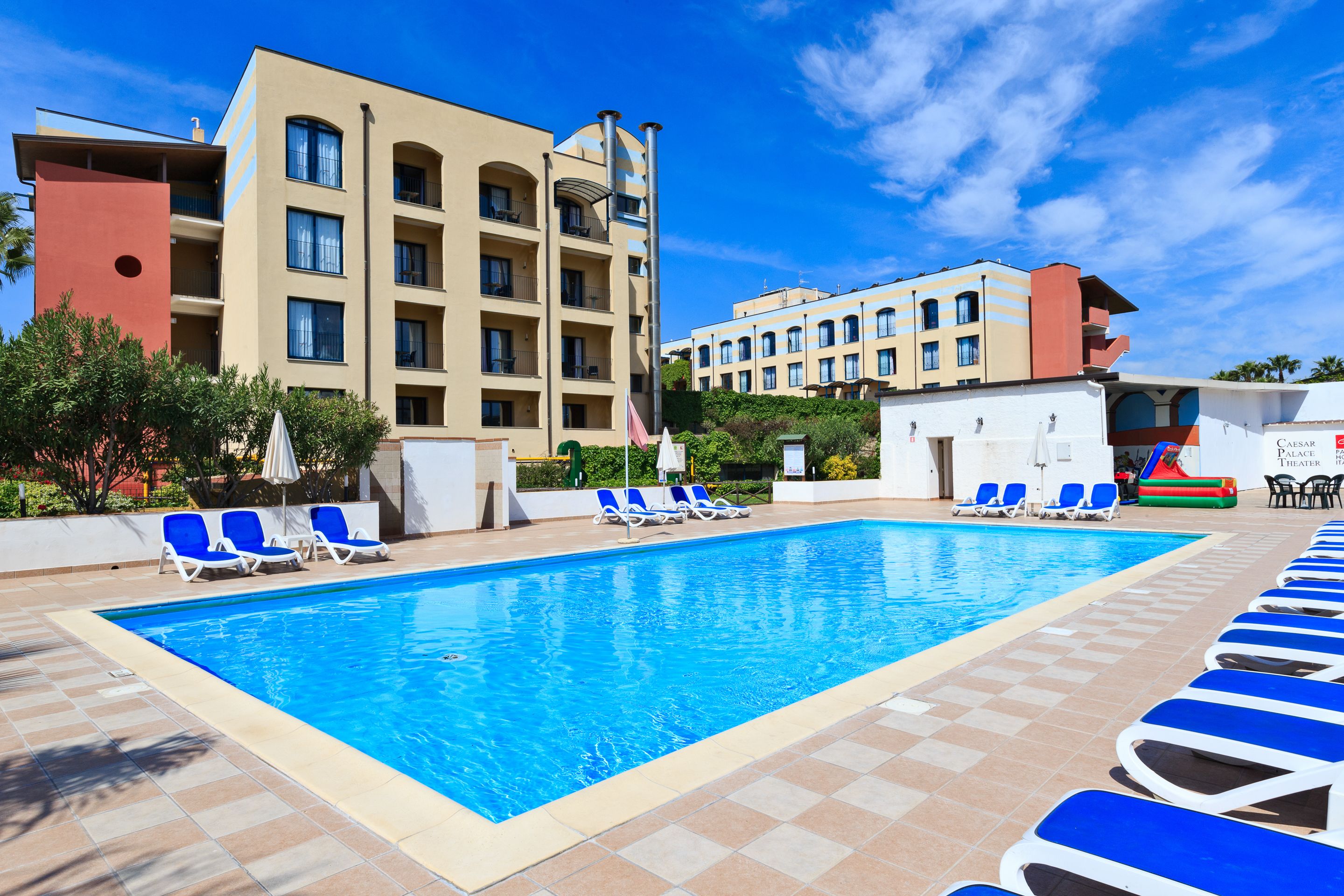 Ein Swimmingpool mit blauem Wasser, umgeben von Liegestühlen und einem Gebäude mit mehreren Balkonen.