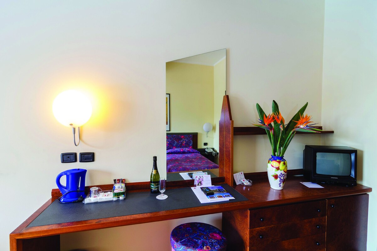A hotel room with a desk, a mirror, a TV, a vase with flowers, a bottle, and a coffee maker.
