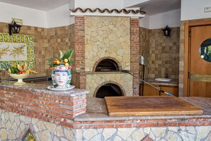 A kitchen with a stone fireplace, a fruit bowl, and a wooden cutting board. - Hotel Caesar Palace (Giardini Naxos)