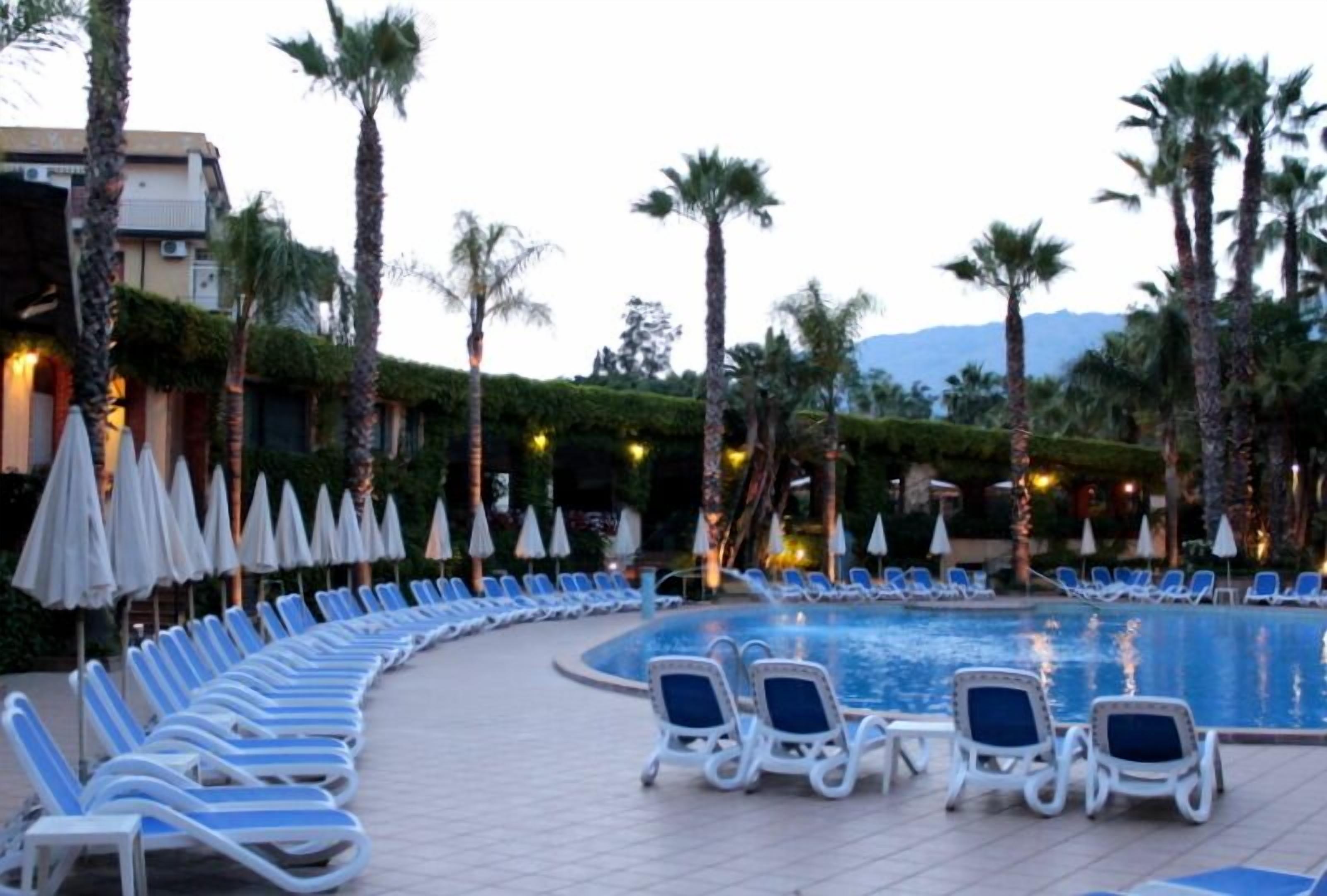 Area piscina con sedie a sdraio e ombrelloni, circondata da palme e un edificio con vista sulle montagne sullo sfondo.