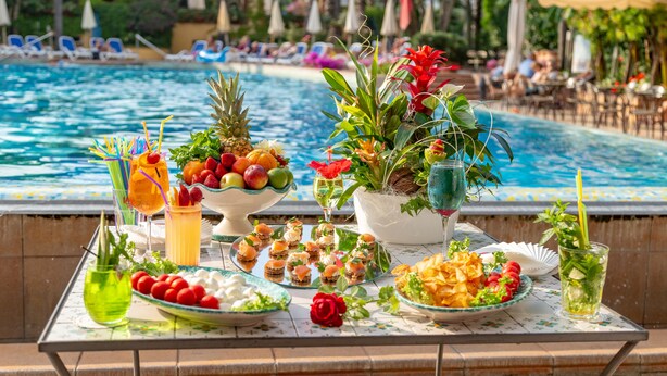 Una mesa junto a la piscina con diversos aperitivos, bebidas y un centro de mesa decorativo.