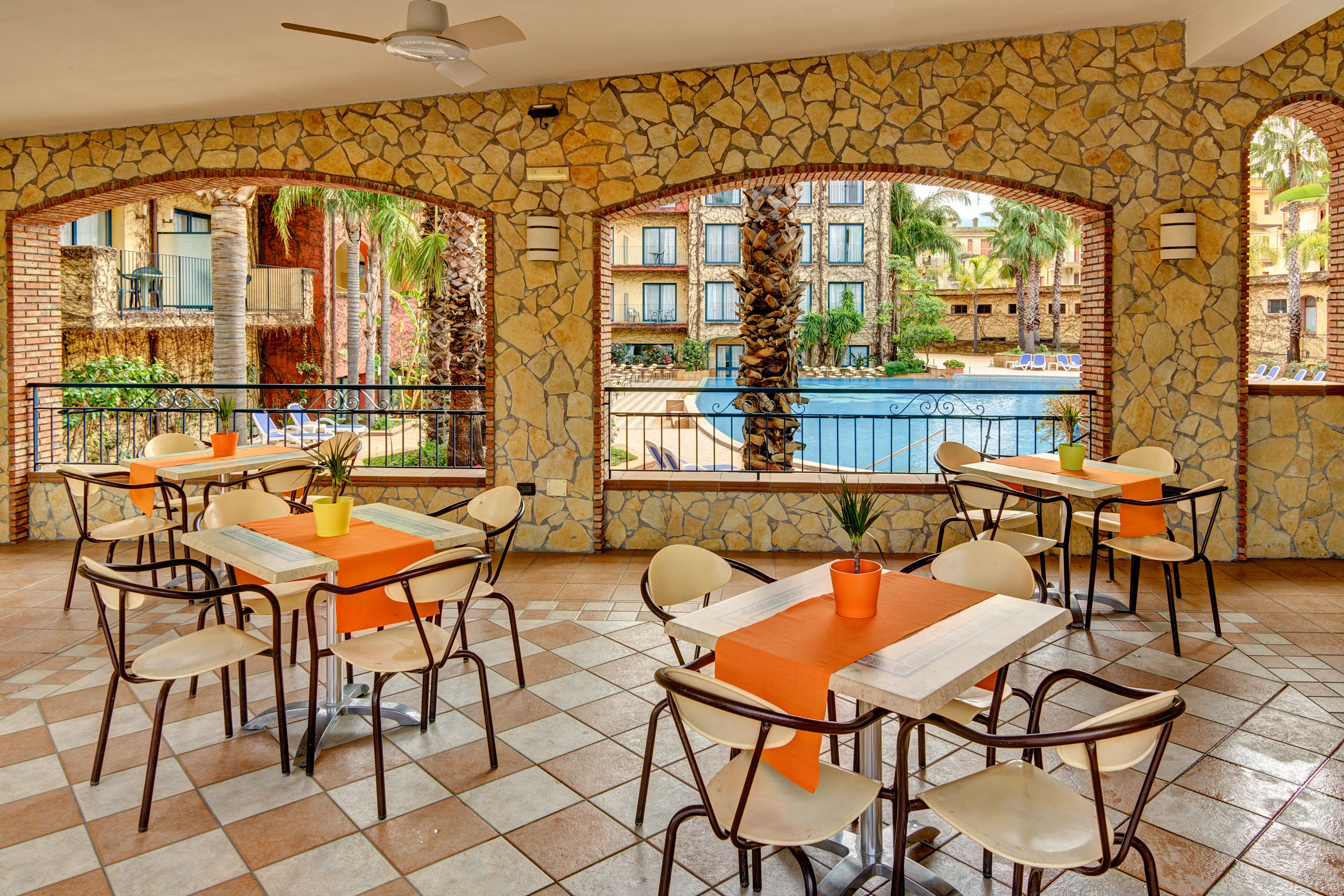 Un restaurante con terraza, paredes de piedra y vistas a una piscina y edificios.