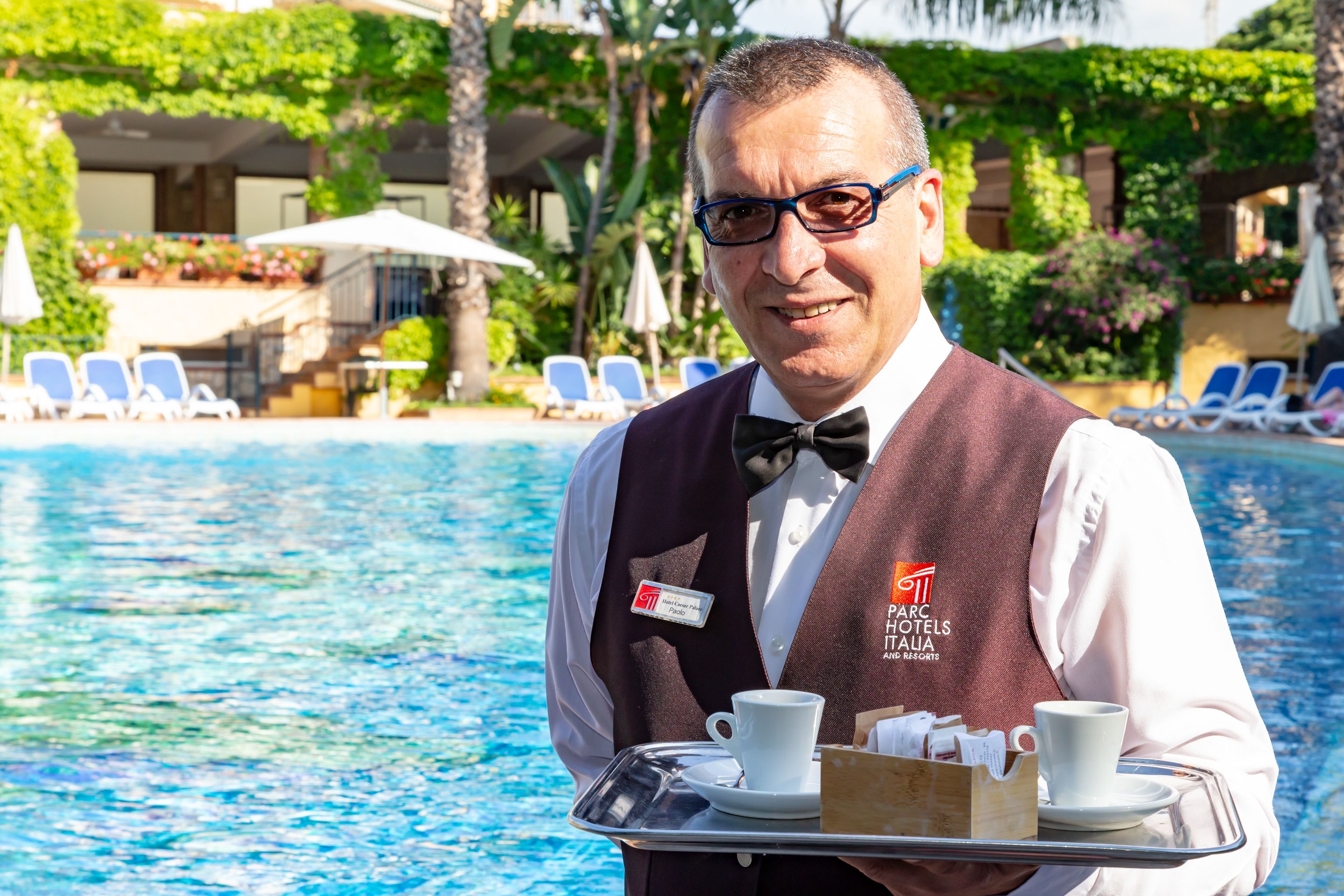 Un miembro del personal de hotel sostiene una bandeja con dos tazas y una caja de bombones junto a una piscina.