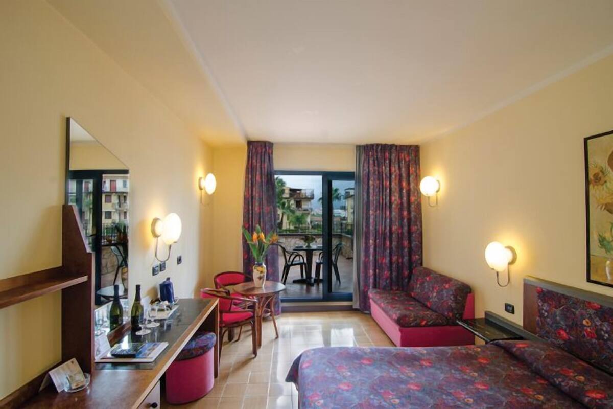 Une chambre d’hôtel comprenant un lit, un bureau, une chaise, une petite table et un balcon avec des rideaux.