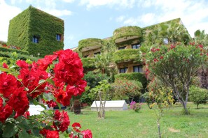A building with a unique green, ivy-covered exterior and a garden with red flowers in the foreground. - Hotel Caesar Palace (Giardini Naxos)