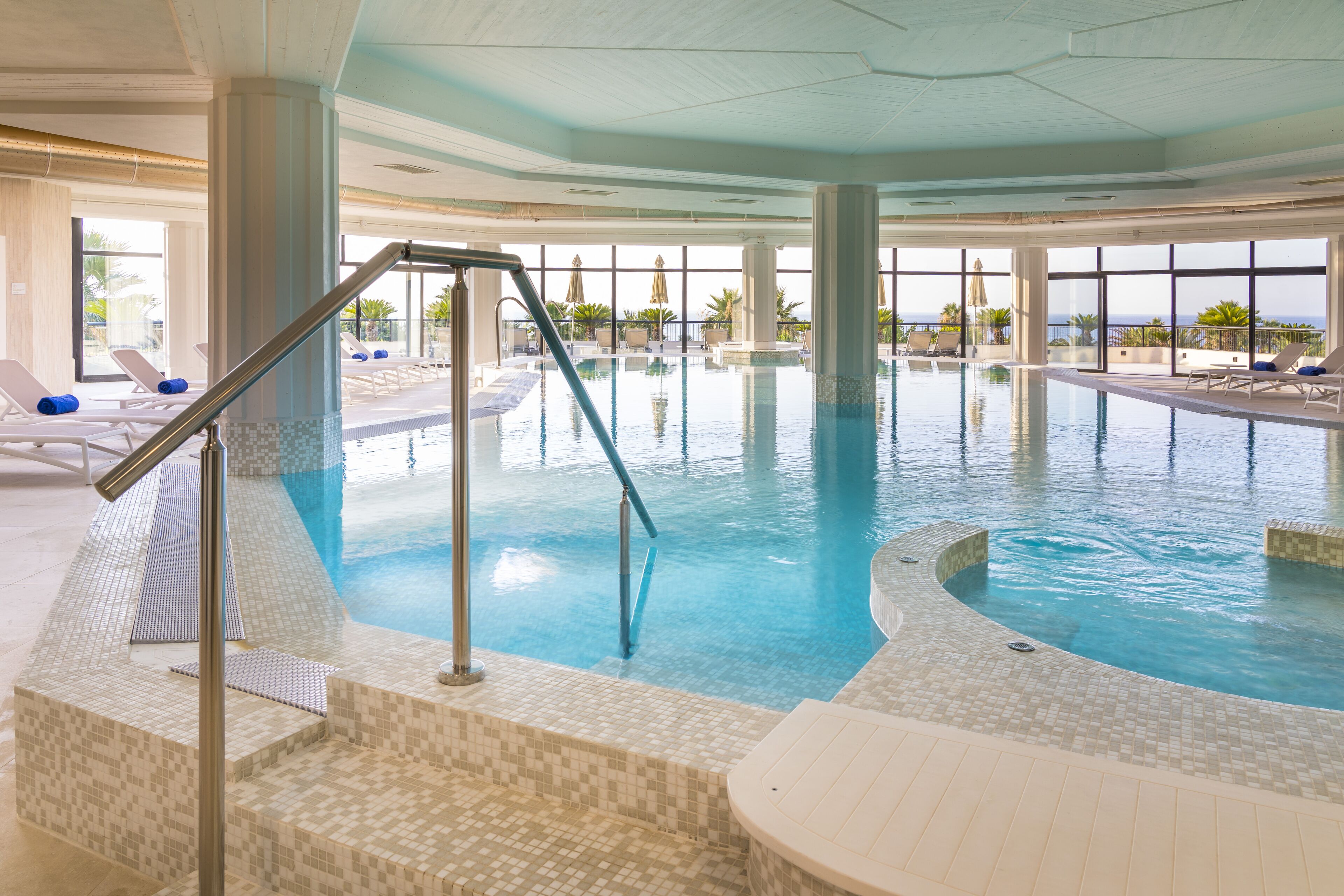 An indoor swimming pool with tiled steps, lounge chairs, and large windows offering a view of the ocean.
