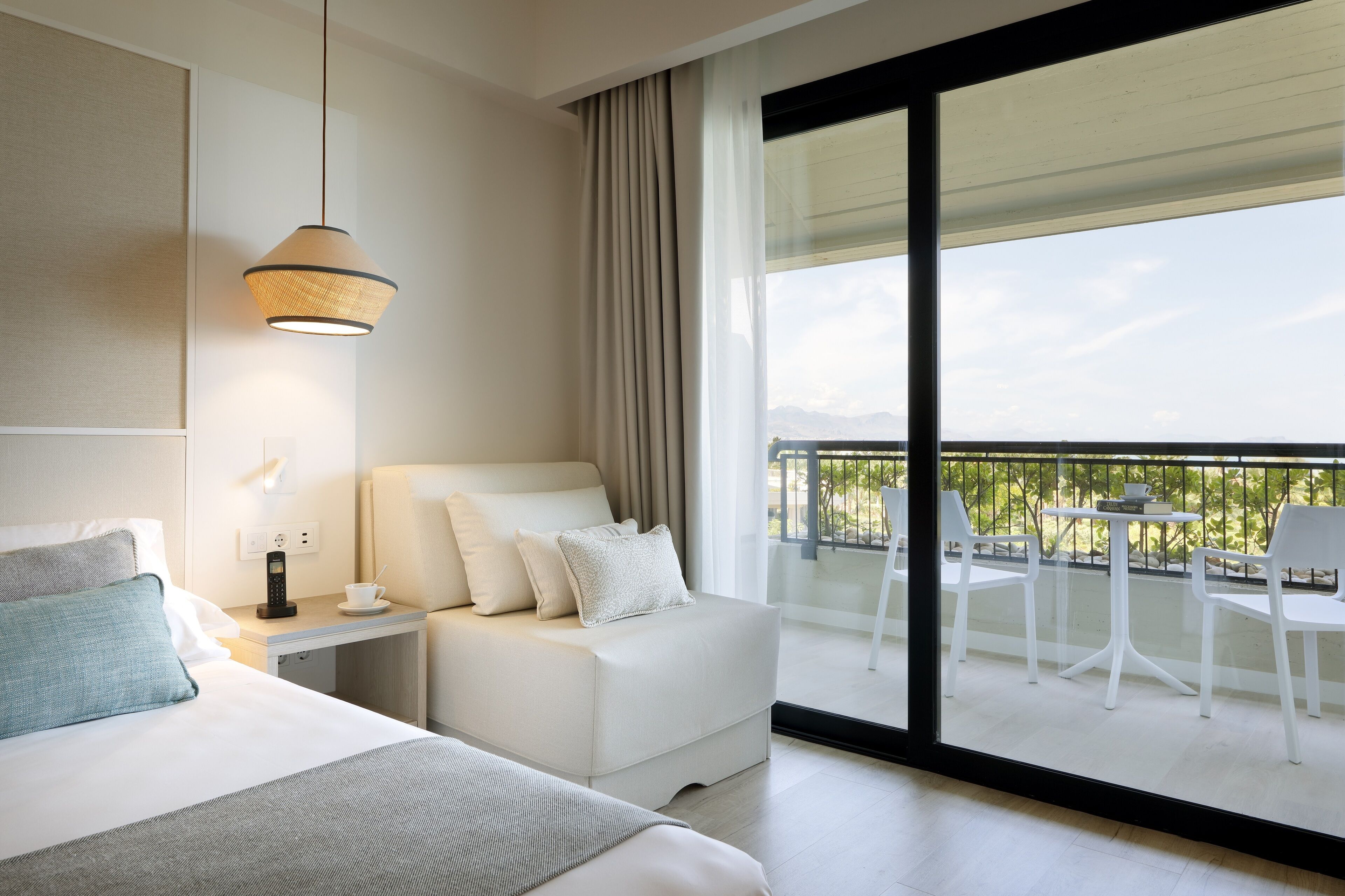 A modern hotel room with a bed, bedside table, and a balcony with outdoor seating.