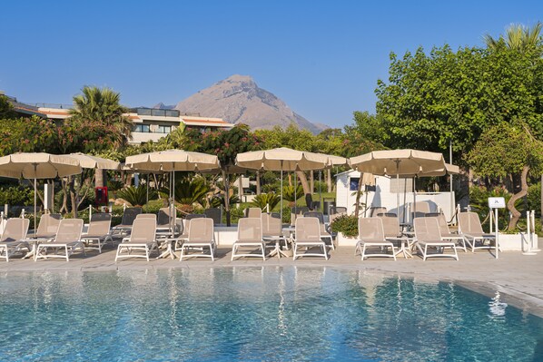 Zona de piscina con sillas de descanso y sombrillas, una montaña al fondo y cielo despejado.