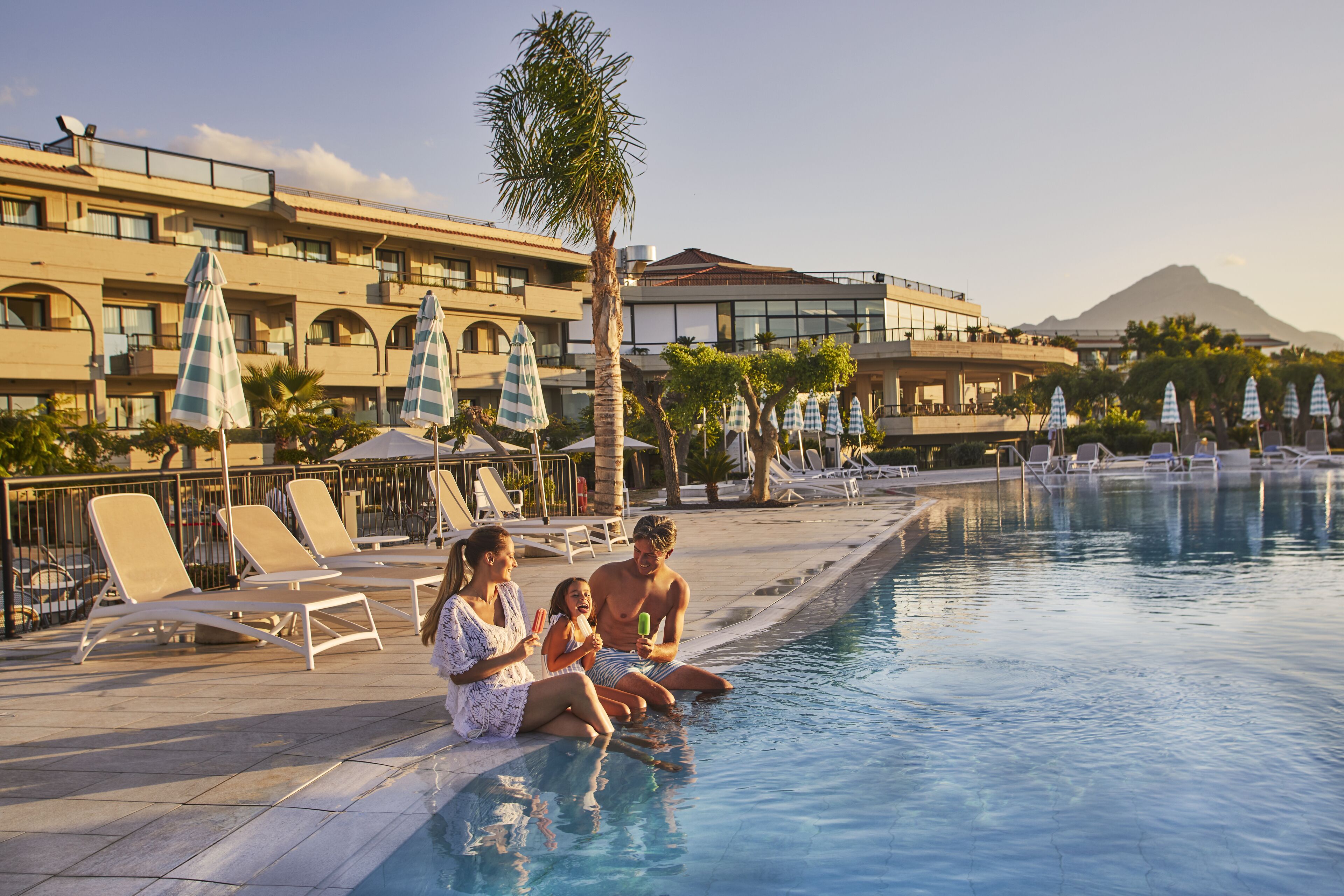 A swimming pool area with lounge chairs and umbrellas, a palm tree, and a building with balconies in the background.