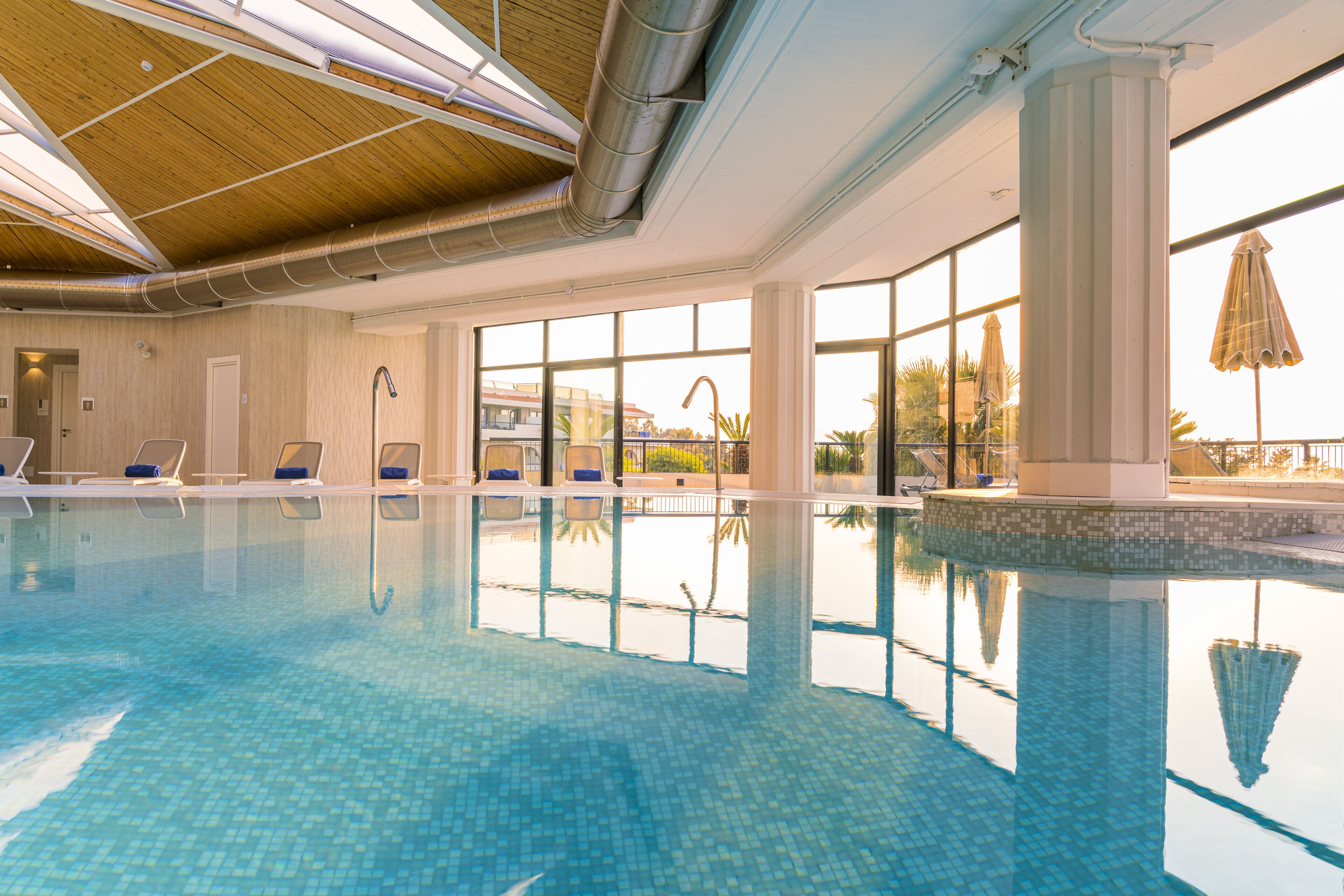 Una piscina interior con agua azul cristalina, sillas de descanso y amplias ventanas que ofrecen vistas a palmeras y un edificio.