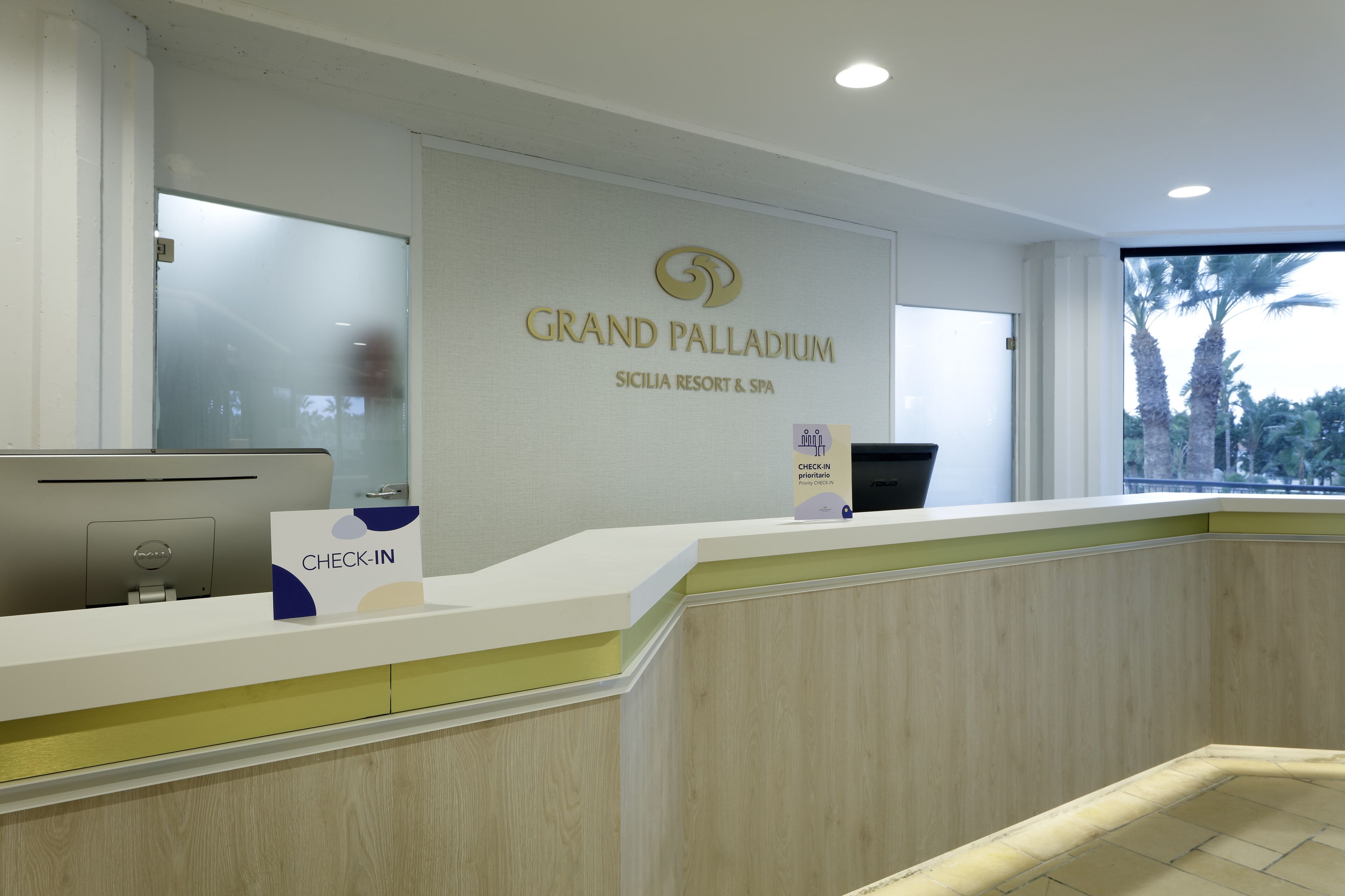 Una reception al Grand Palladium, con un cartello che indica 'Check-In', un computer e una finestra vista palme.