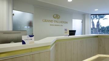 Una reception al Grand Palladium, con un cartello che indica 'Check-In', un computer e una finestra vista palme.