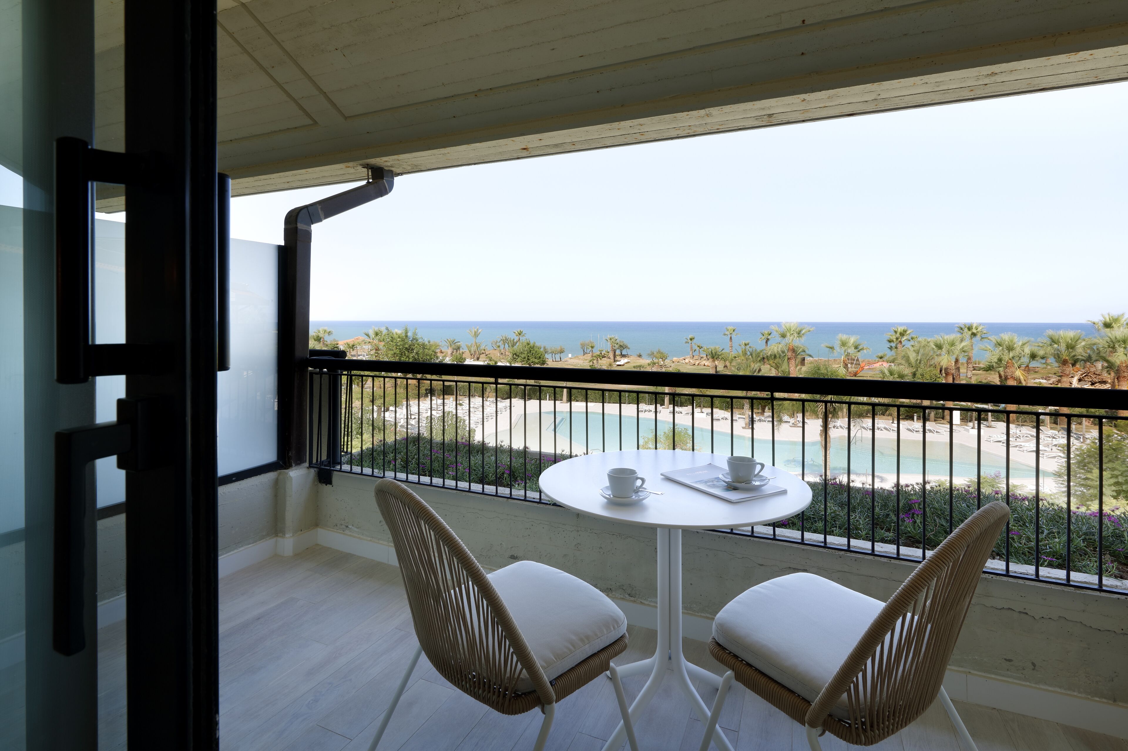 A balcony with a white table and two wicker chairs, overlooking a beach with palm trees and a clear view of the ocean.