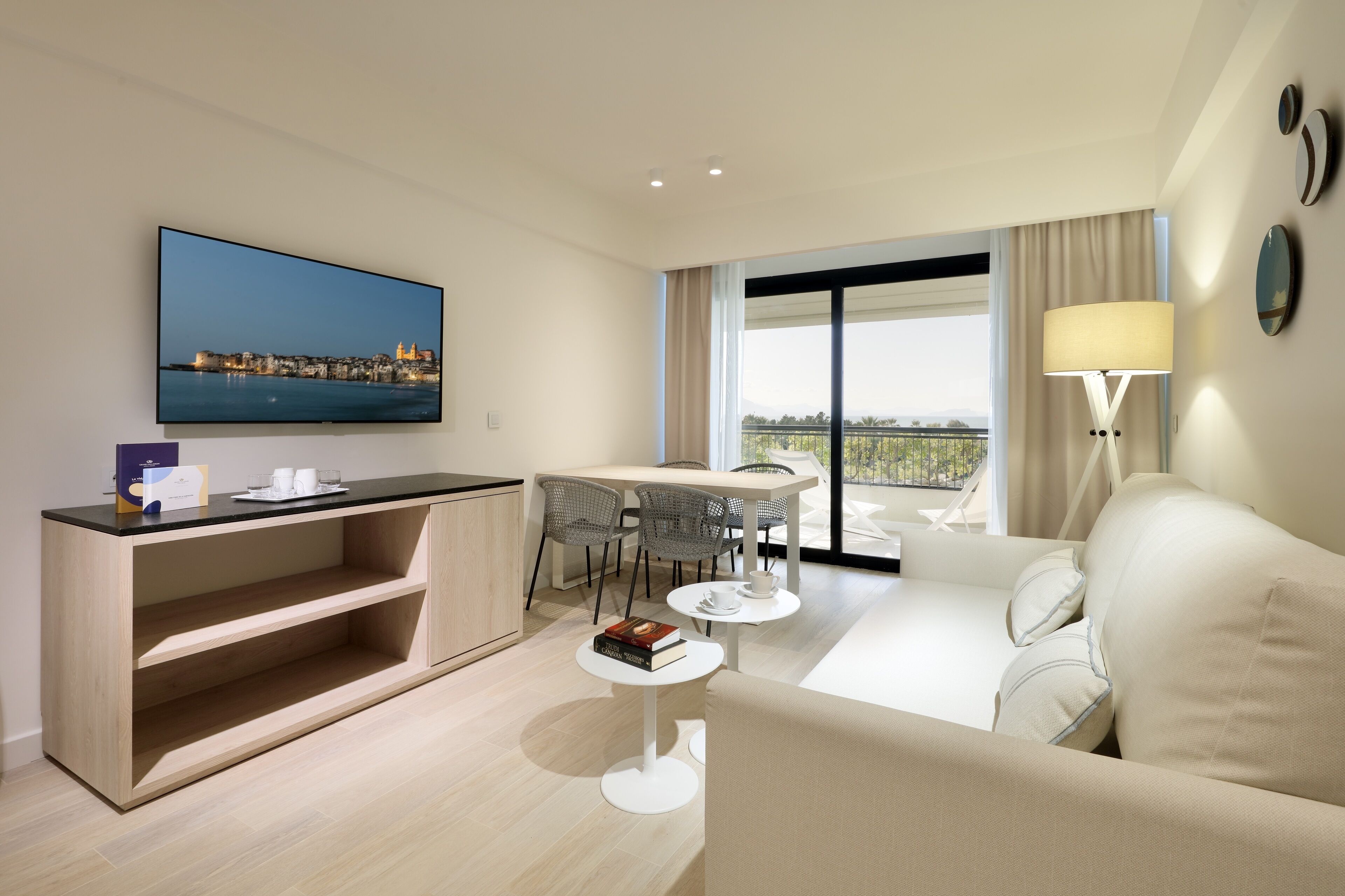 A modern hotel room with a flat-screen TV, a dining area with a table and chairs, a sofa, a small coffee table, and a large window with a view.