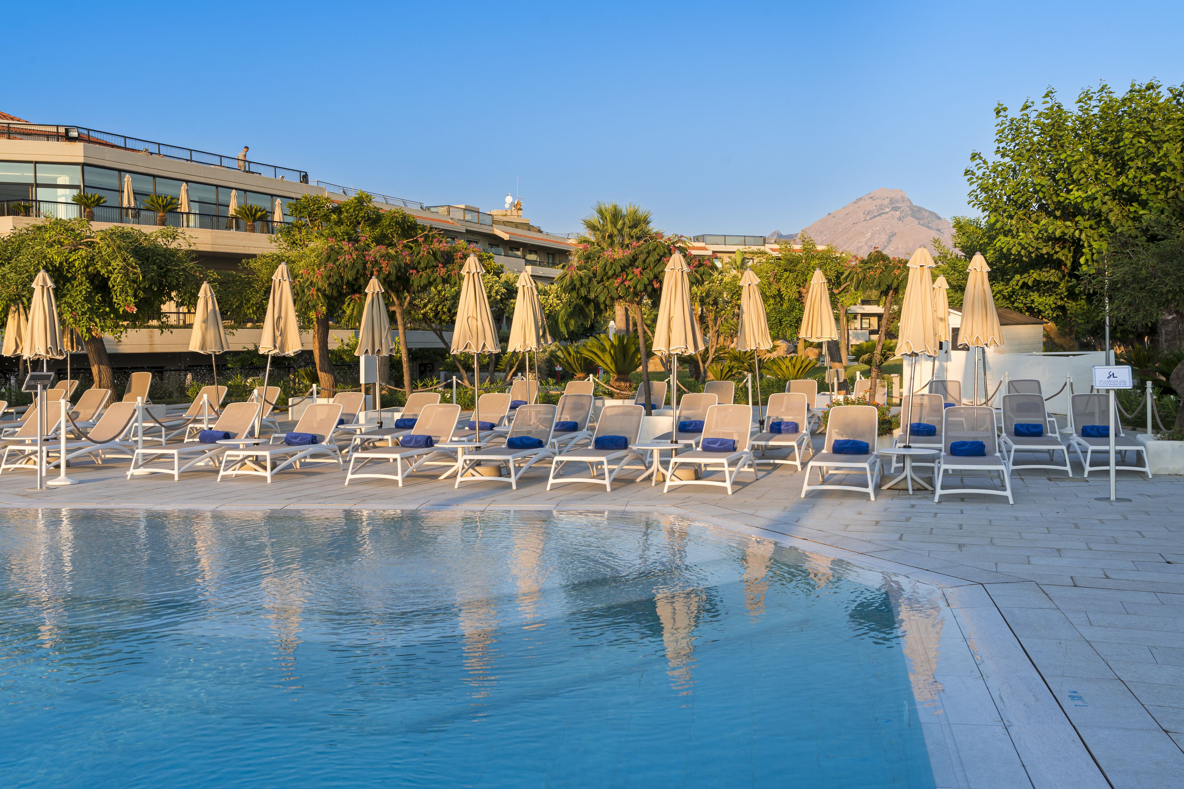 Zona piscina con lettini e ombrelloni, cielo sereno e una montagna sullo sfondo.