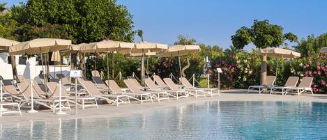 A poolside area with lounge chairs and umbrellas, surrounded by lush greenery and flowering plants.