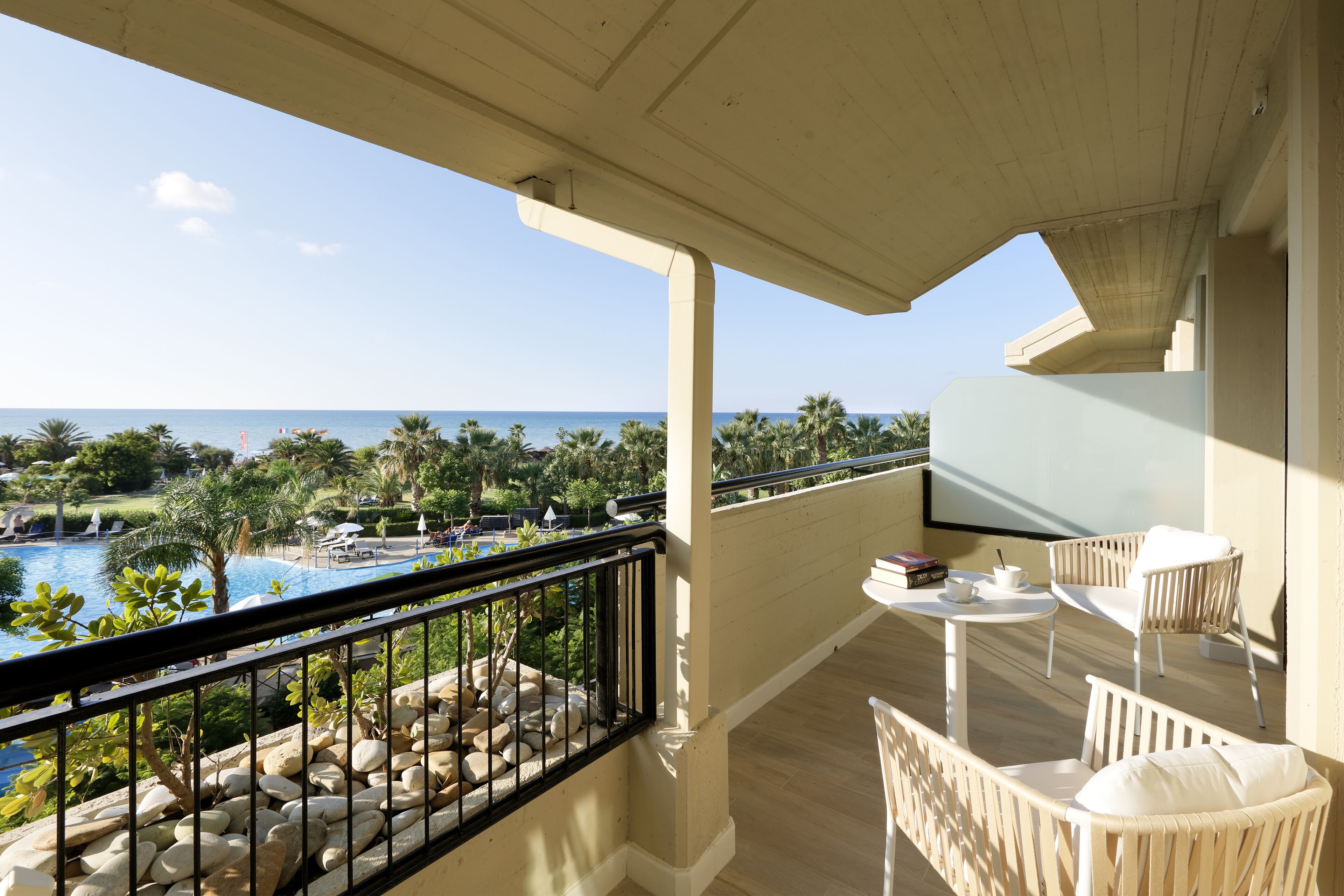 A balcony with a view of a pool and palm trees, a table with chairs, and a small book and cup on the table.