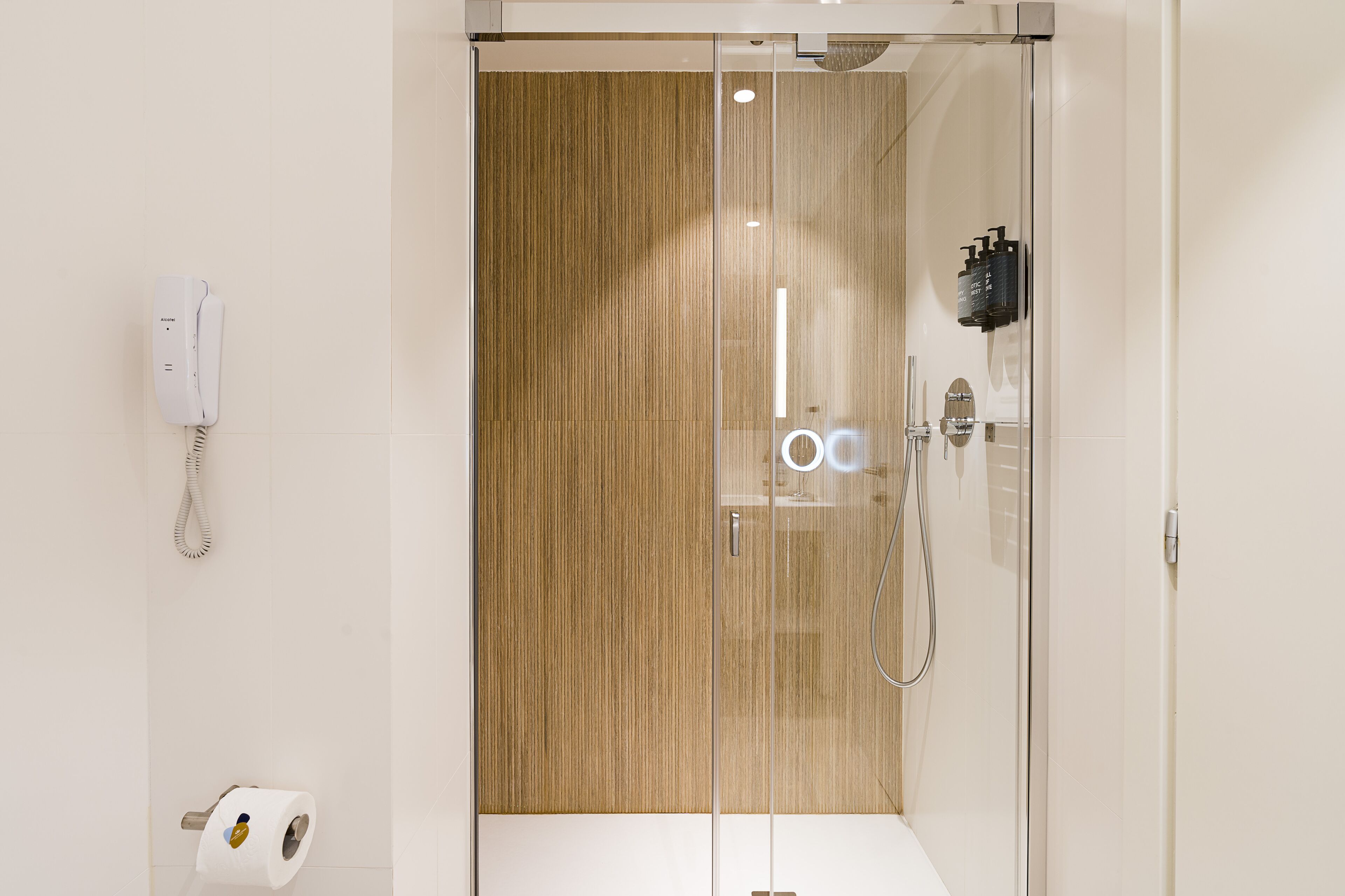 A modern bathroom with a glass shower enclosure, a toilet paper dispenser, and a telephone mounted on the wall.