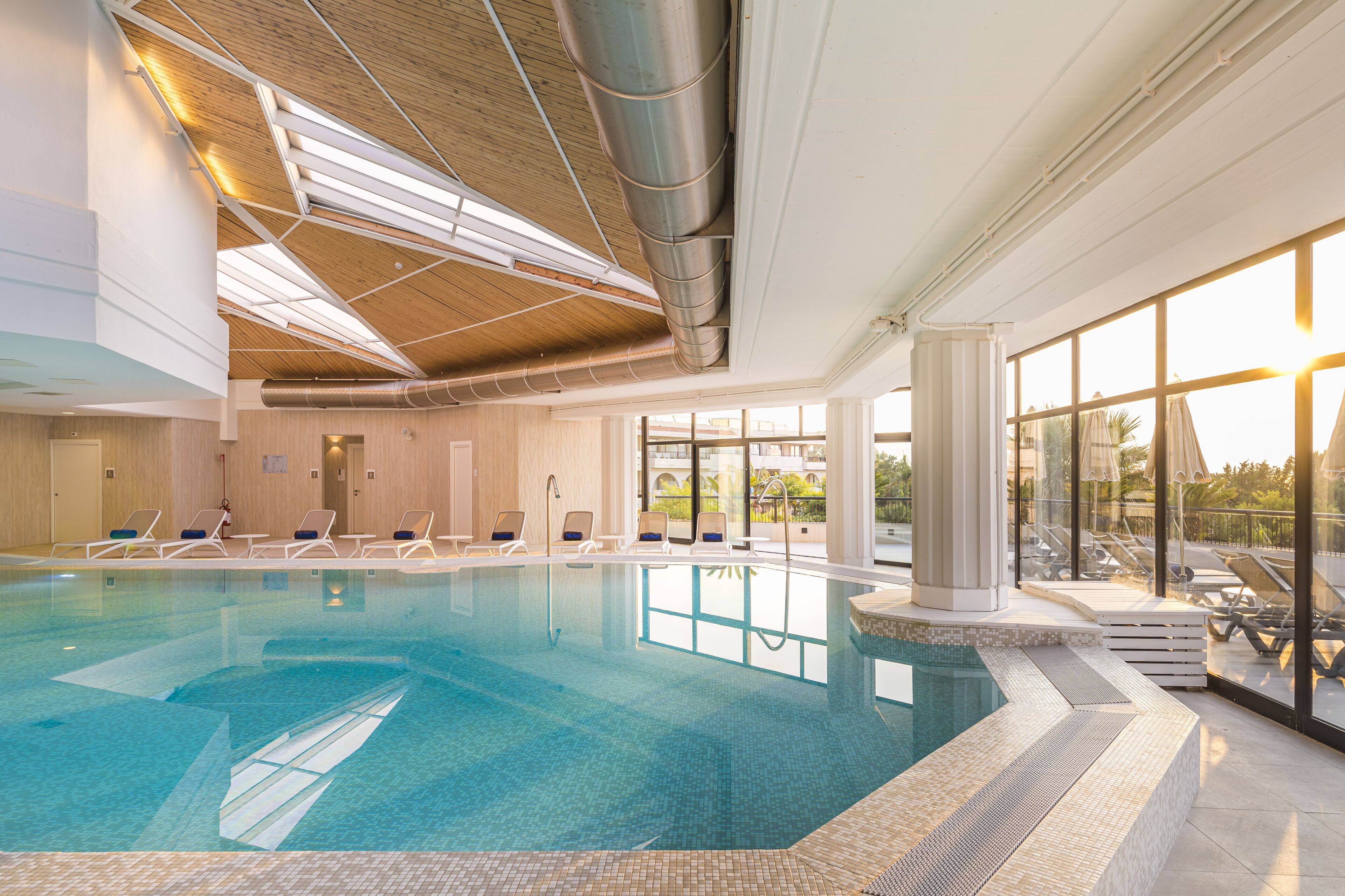An indoor swimming pool with clear water, surrounded by lounge chairs and a view of the outdoors through large windows.