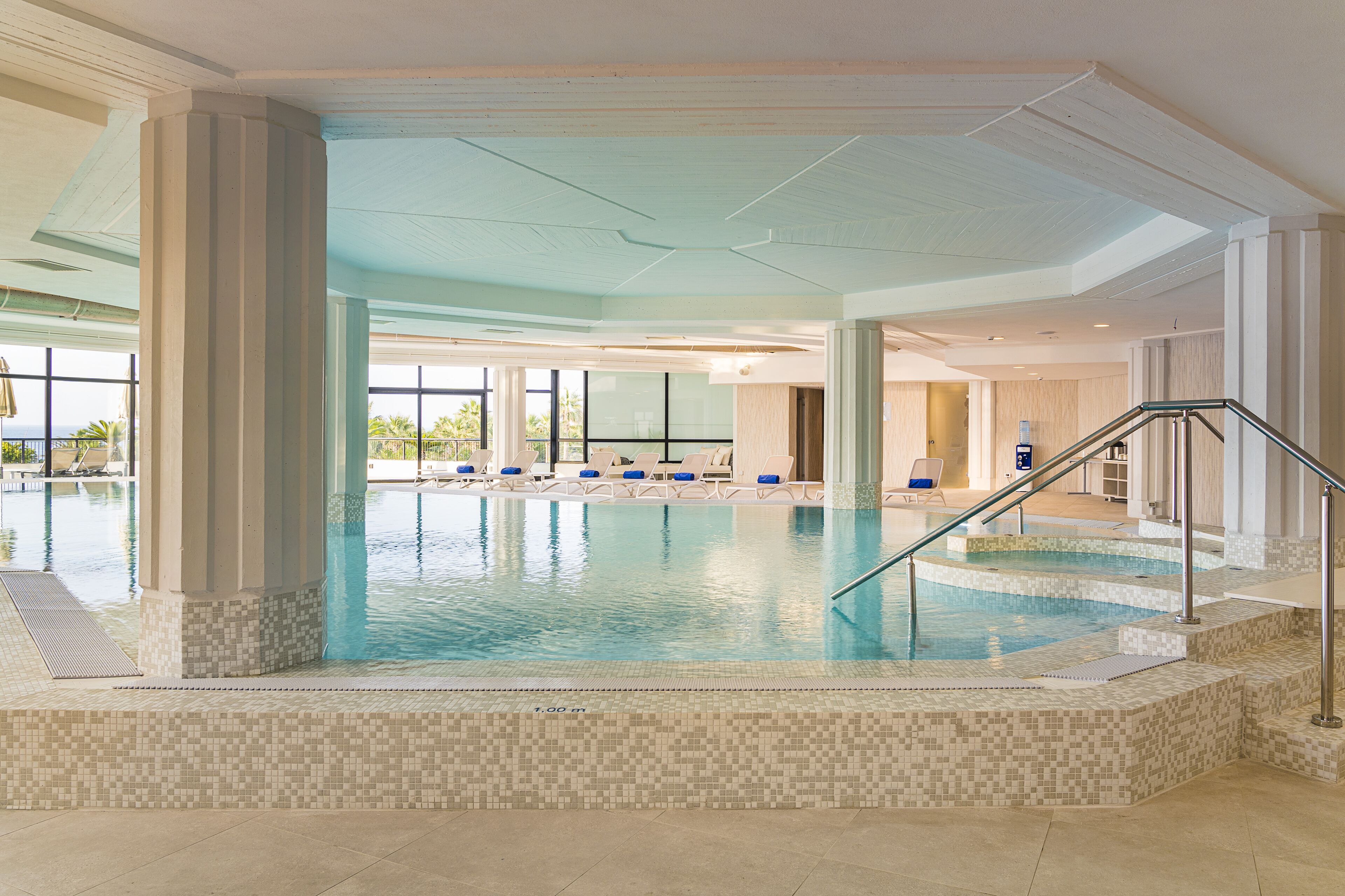 Une piscine intérieure aux murs et au sol carrelés, entourée de chaises longues et d’un escalier menant aux niveaux supérieurs.