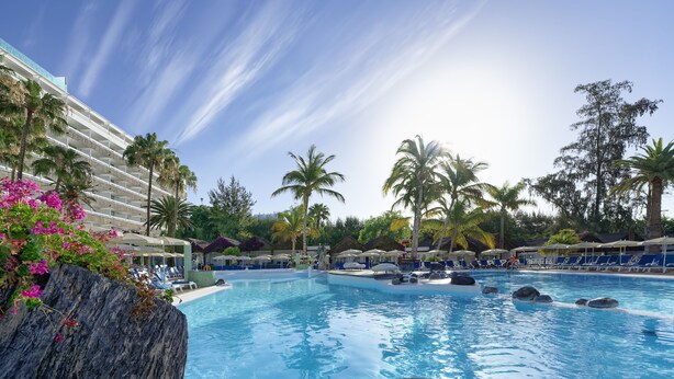 Área de piscina de un resort con agua azul cristalina, sillas de descanso, sombrillas, palmeras y un edificio moderno con balcones.