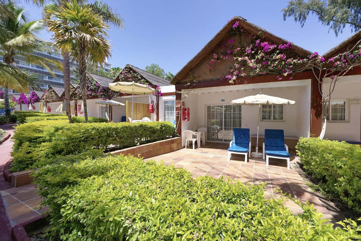 A resort with a patio area featuring two lounge chairs, a covered seating area, and a well-maintained garden with hedges and palm trees.