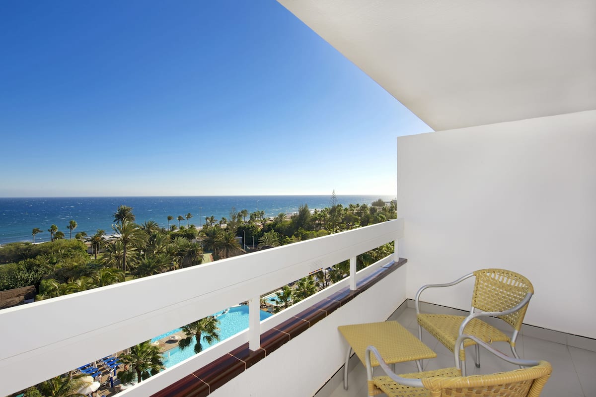 A balcony with a view of a pool and ocean, featuring wicker furniture and a white railing.