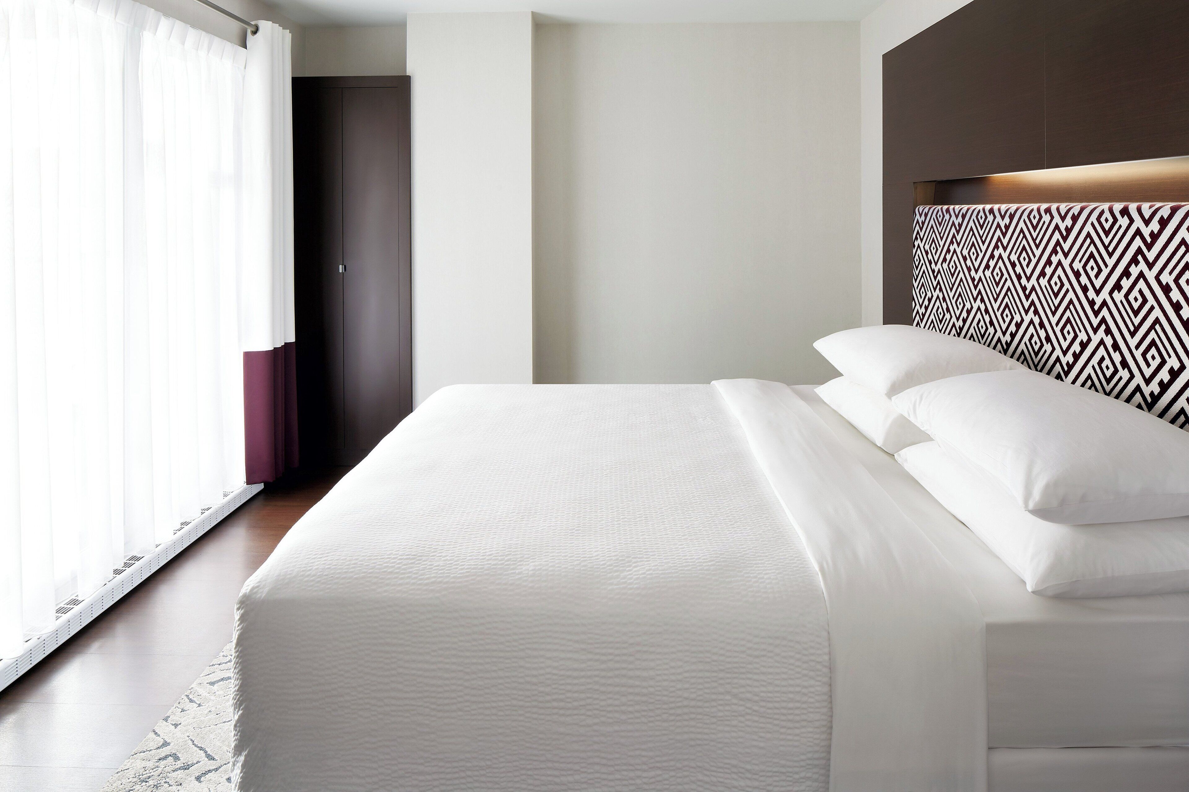 A modern hotel room with a large bed, a patterned headboard, and a view of the hallway.