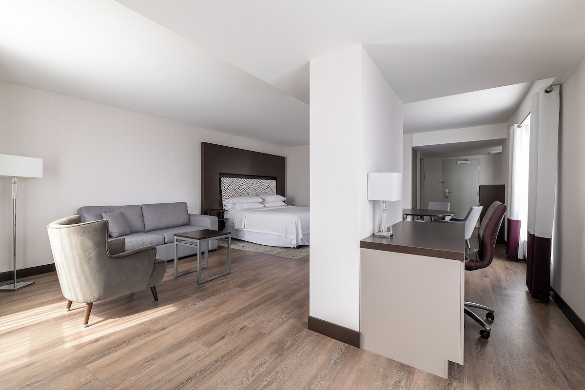A modern hotel room with a grey sofa, a bed with white linens, a desk with a chair, and a small table.