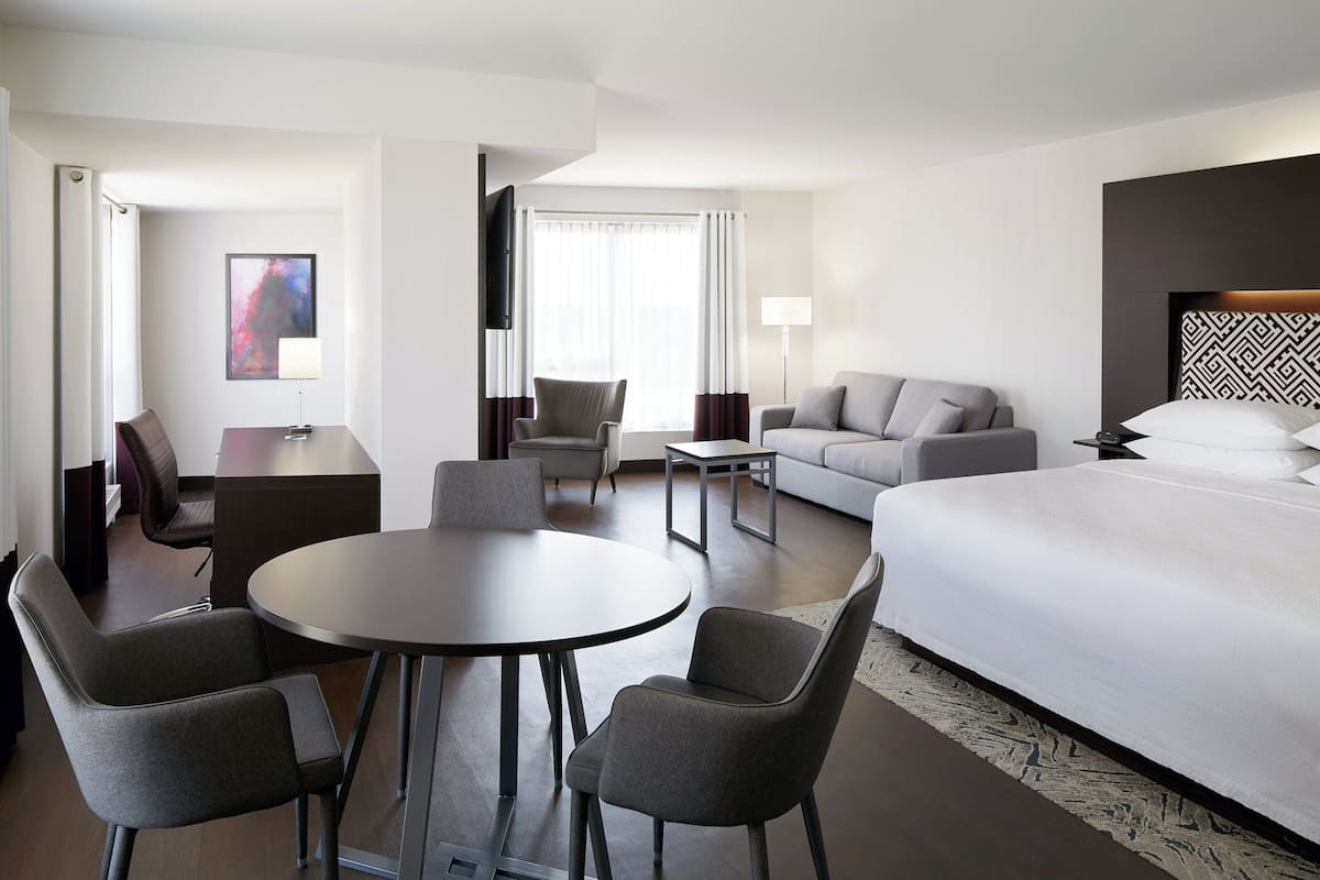 A modern hotel room with a large bed, a sofa, a dining table with chairs, a TV, and a desk.