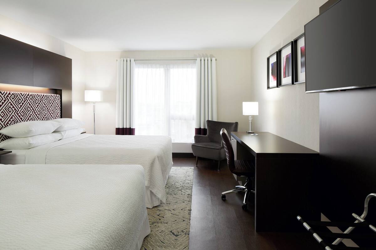 A hotel room with two beds, a desk, a chair, a TV, and a window with curtains.