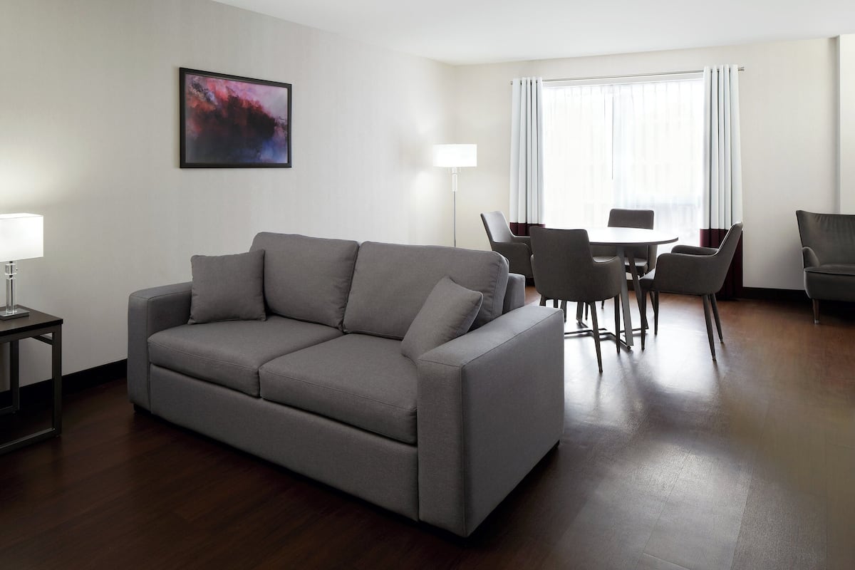A modern living room with a grey sofa, a dining table with chairs, a floor lamp, and a large window with curtains.