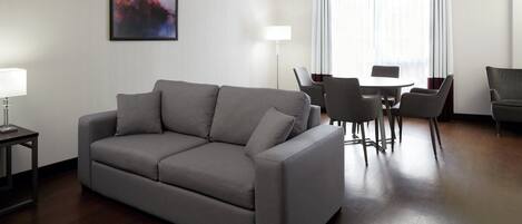 Una sala de estar moderna con un sofá gris, una mesa de comedor con sillas, una lámpara de pie y un gran ventanal con cortinas.