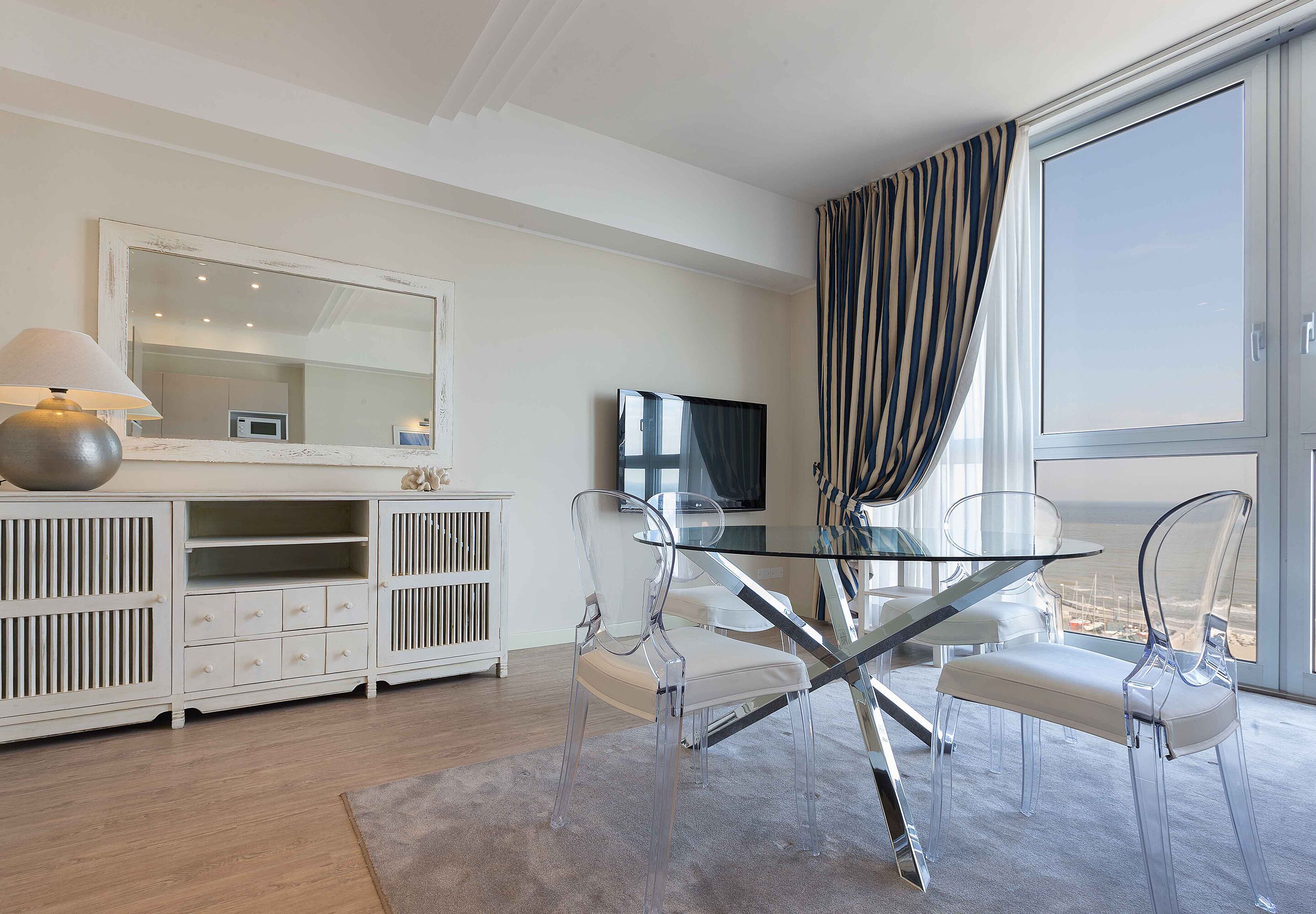 A modern living room with a glass dining table, clear chairs, a white cabinet, a large mirror, and a TV.