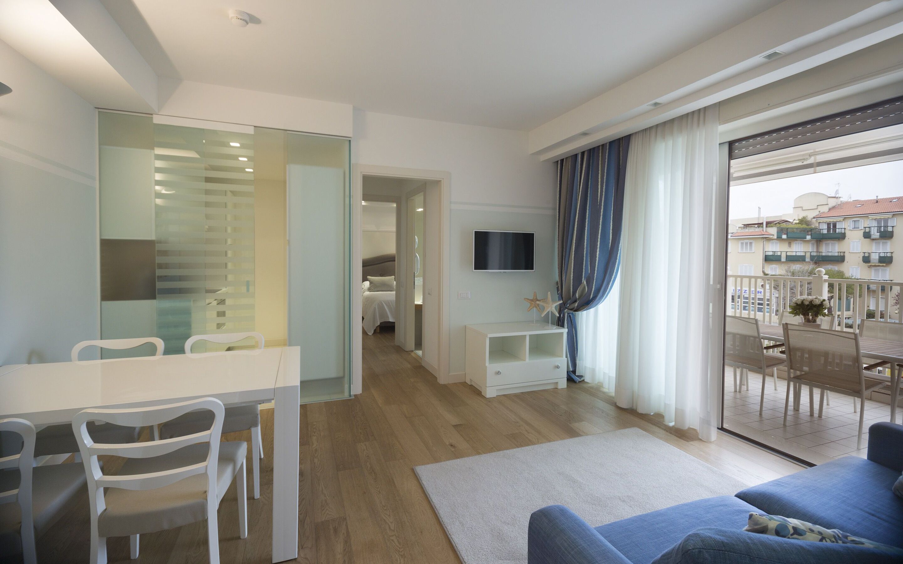 A modern hotel room with a dining area, a TV, and a balcony with a view of buildings.