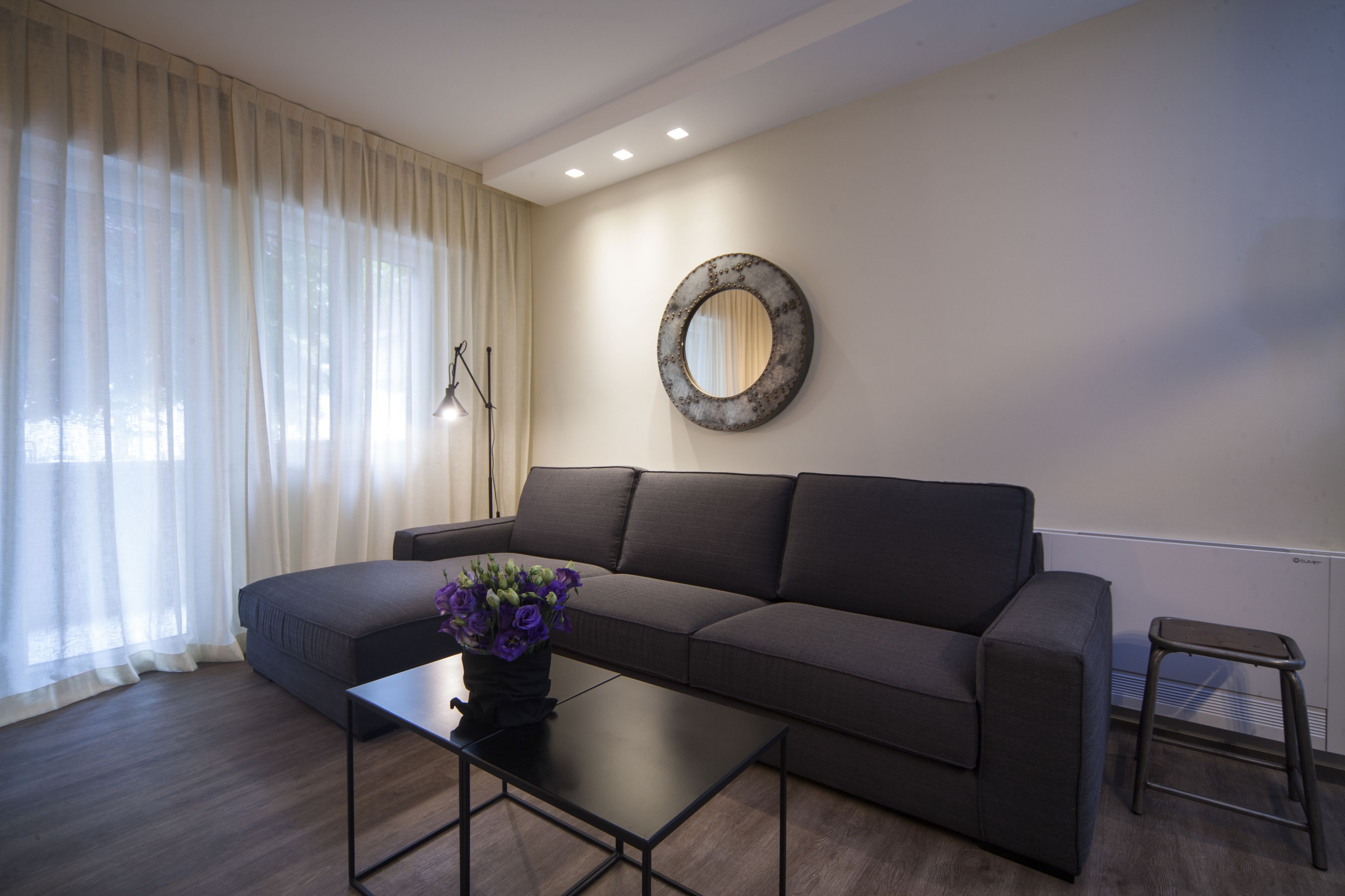 A modern living room with a dark grey sofa, a small black coffee table with a vase of purple flowers, and a round mirror on the wall.