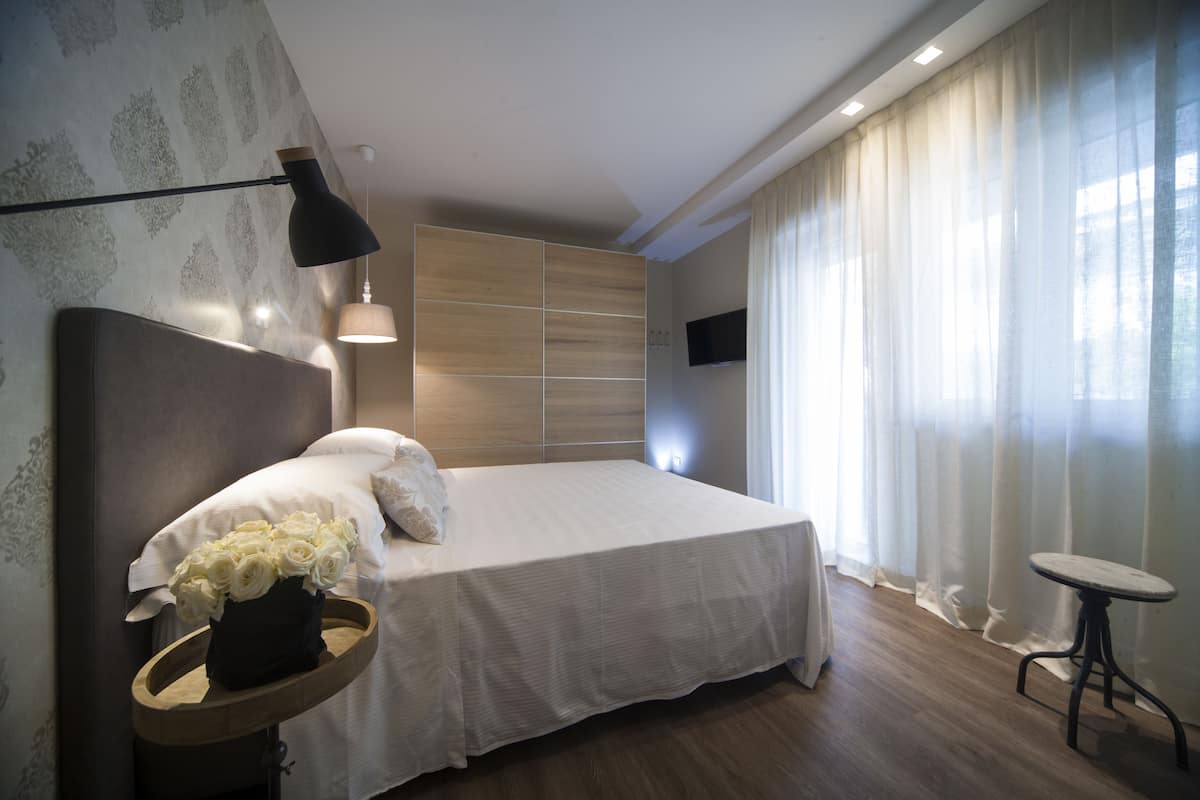 A modern bedroom with a large bed, a bedside table with a vase of flowers, a wall-mounted TV, and a small table by the window.