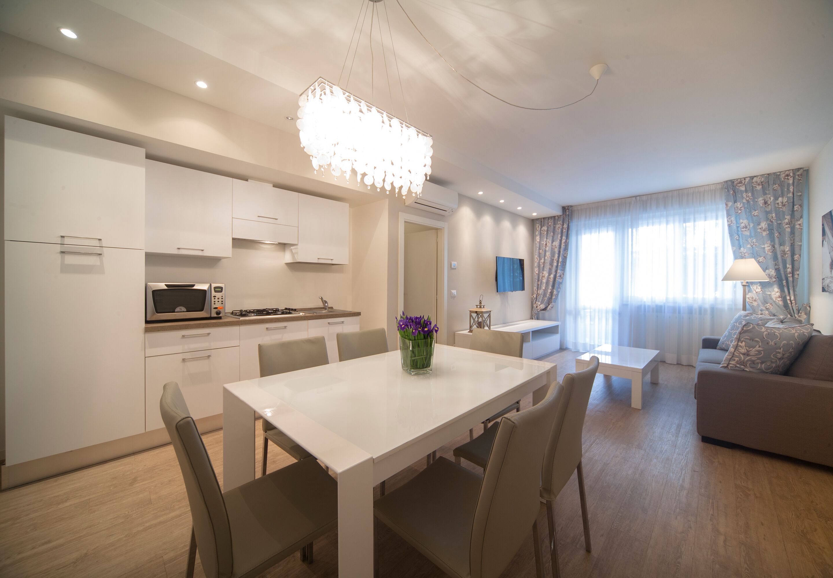 A modern kitchen with white cabinets, a dining table with chairs, a chandelier, and a living area with a sofa and a TV.