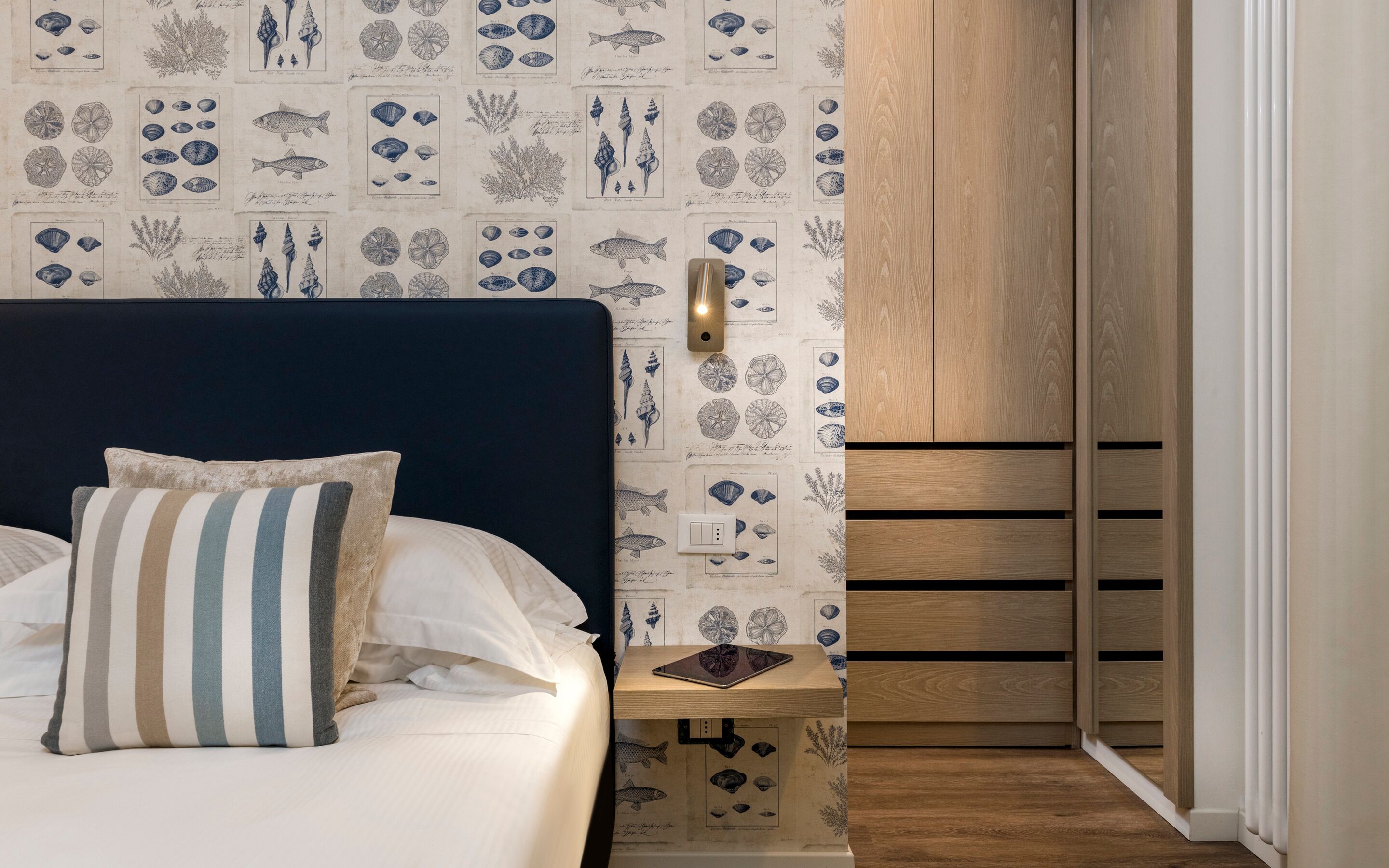 A bedroom with a bed, a bedside table with a tablet, and a wall with marine-themed wallpaper.