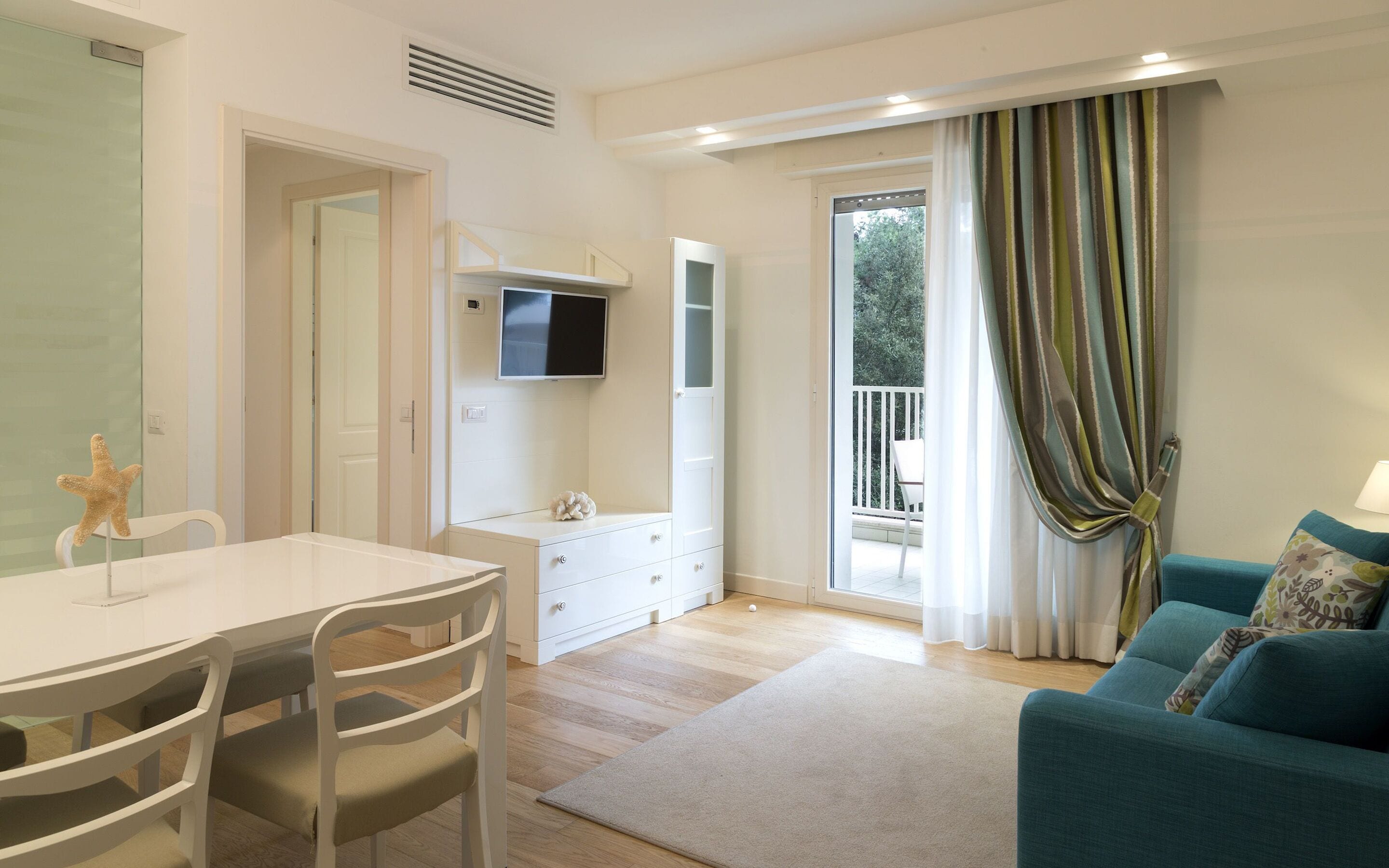 Una sala de estar moderna con una mesa de comedor blanca, sillas, un sofá y un televisor montado en la pared.
