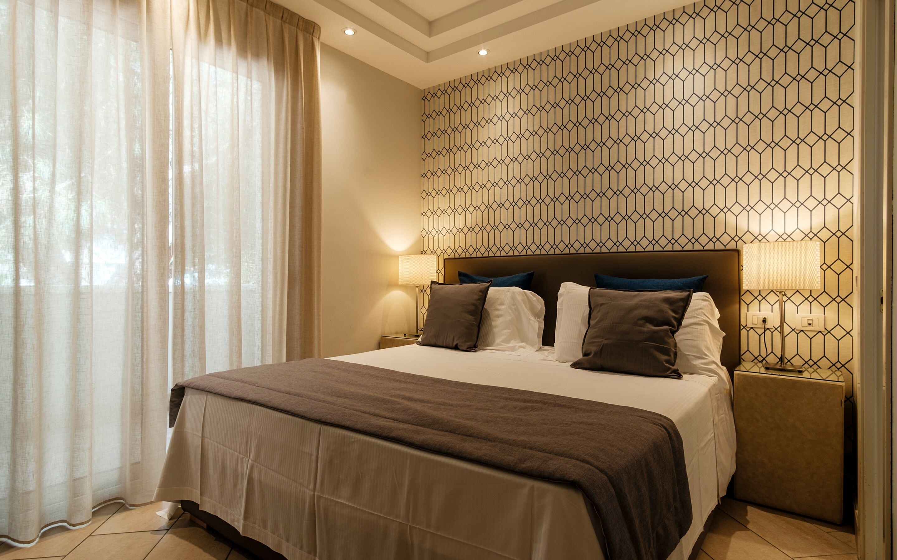 Habitación de hotel con una cama grande, cabecera con diseño geométrico, lámpara de noche y ventana con cortinas traslúcidas.