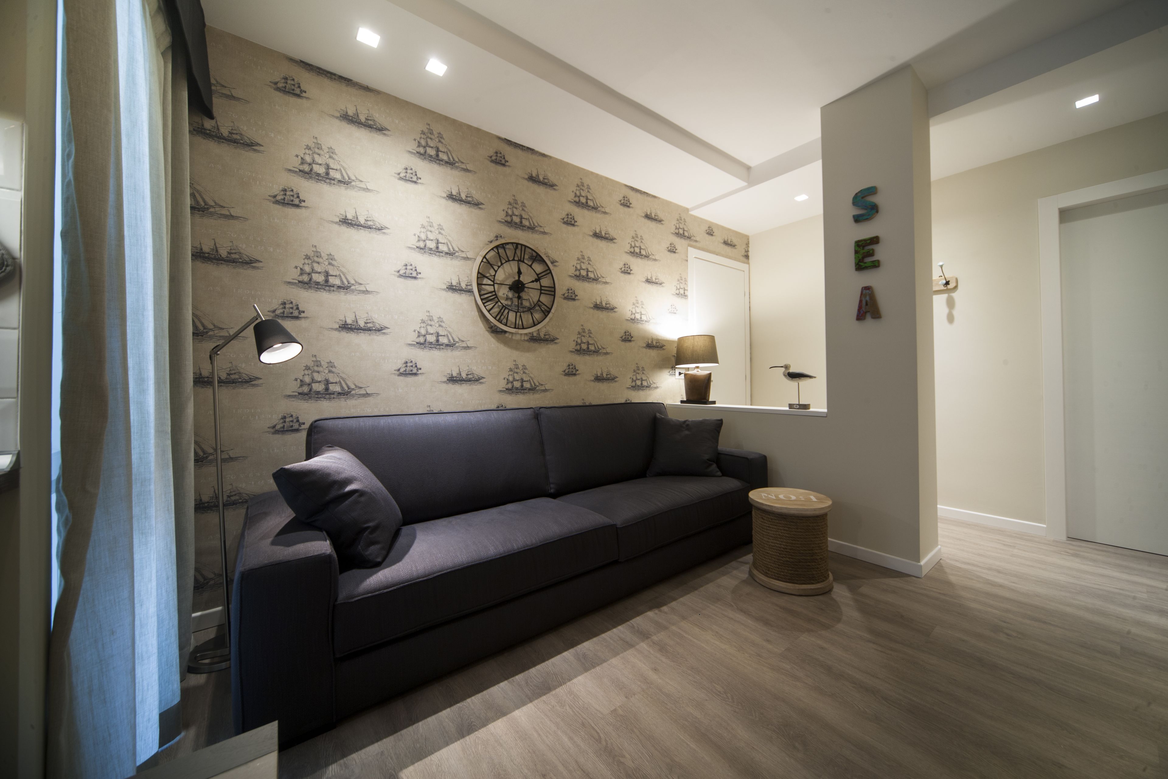 A modern living room with a dark sofa, a patterned wallpaper, a wall clock, and a small lamp.