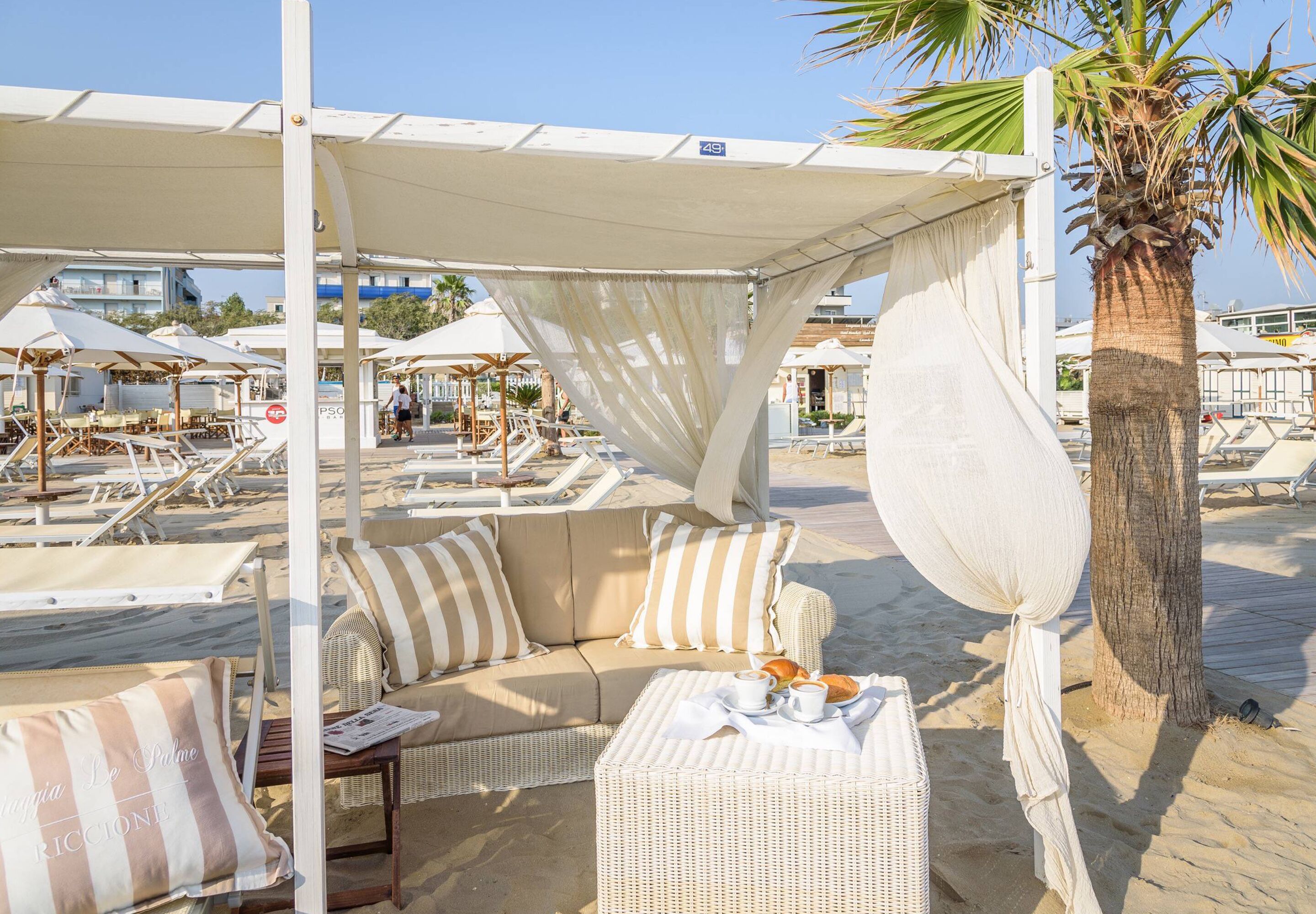 Un espace de détente aménagé sur la plage, avec un canapé et une petite table, un palmier et des parasols en arrière-plan.