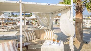 Un espace de détente aménagé sur la plage, avec un canapé et une petite table, un palmier et des parasols en arrière-plan.