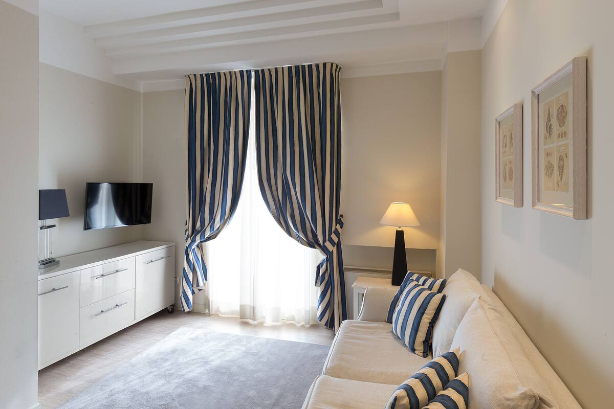 A modern hotel room with a large window, striped curtains, a sofa with striped cushions, a flat-screen TV, a bedside table with a lamp, and framed artwork on the wall.