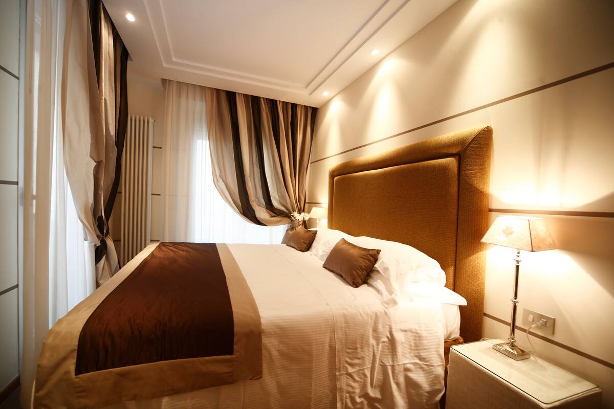 A hotel room with a large bed, a nightstand with a lamp, and a view of the city through the window.